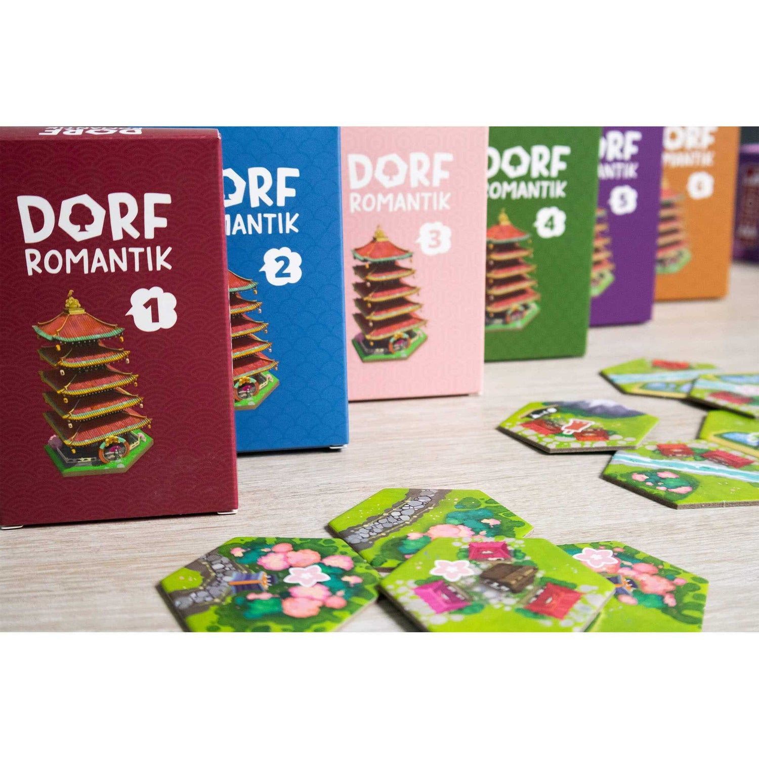 Kartenspielboxen in Rot, Blau, Rosa, Grün, Lila und Orange. Sechseckige Kacheln sind davor verstreut. Auf jeder Box befindet sich eine Pagode.