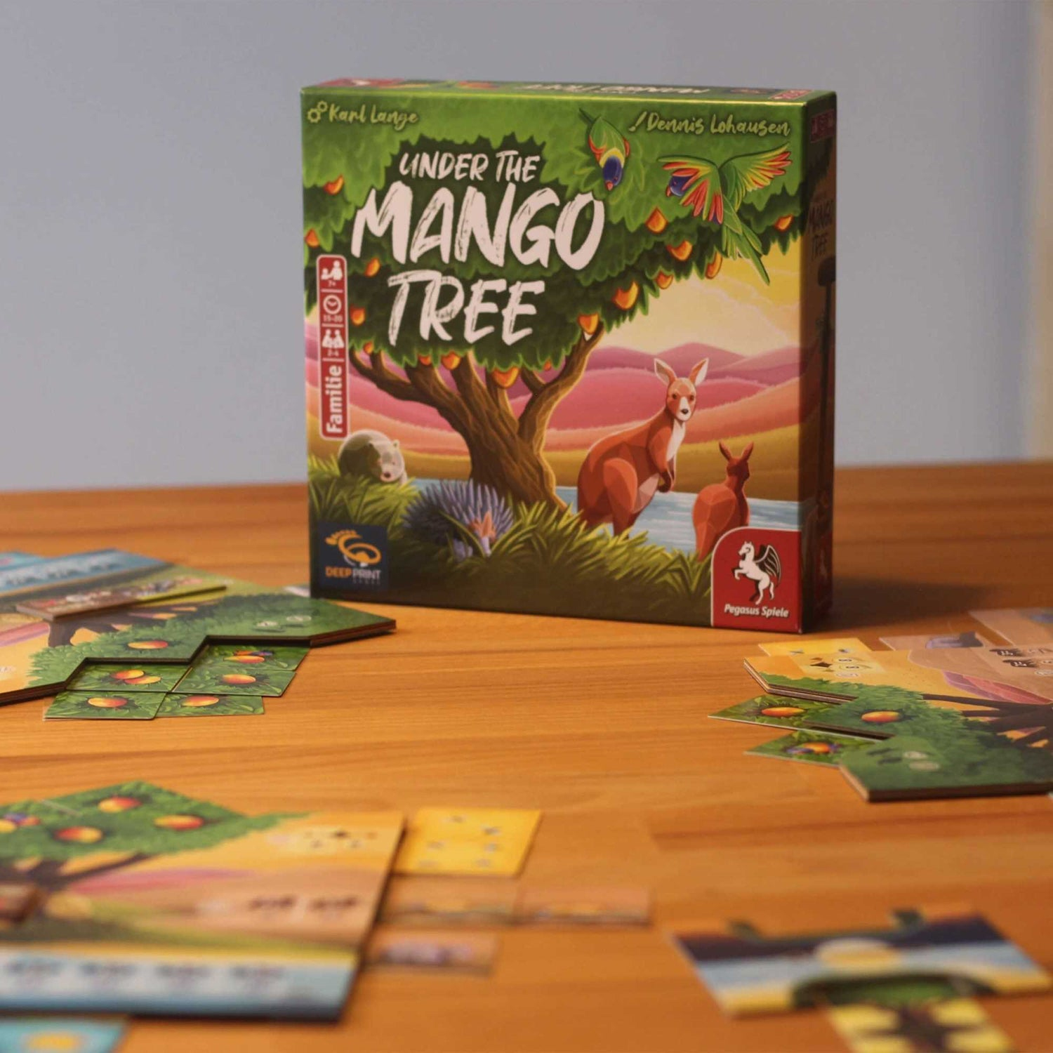 Eine Spieleschachtel mit dem Titel 'Under the Mango Tree' mit Känguru-Illustration und Spielsteinen auf einem Holztisch.