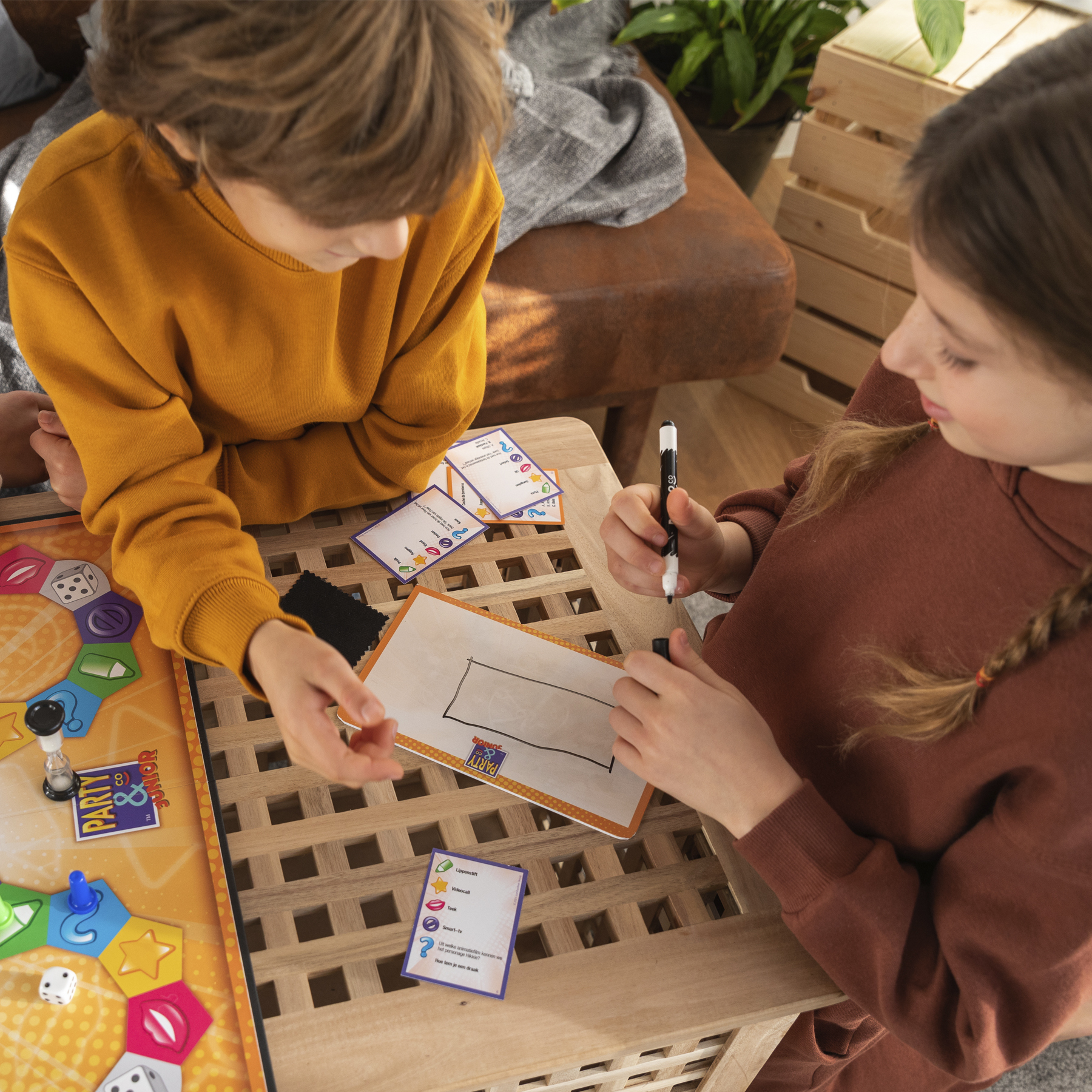 Dos niños jugando un juego de mesa. Uno dibuja en una pizarra, el otro mira cartas del juego. Un dado y un temporizador están en el tablero.