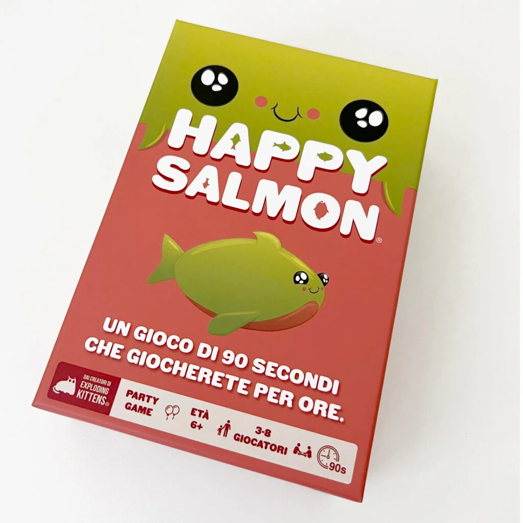 Eine Spielbox von 'Happy Salmon'. Roter Hintergrund mit einem Cartoon-Lachs. Grüner Deckel mit Augen und dem Spieltitel. Weißer Hintergrund.
