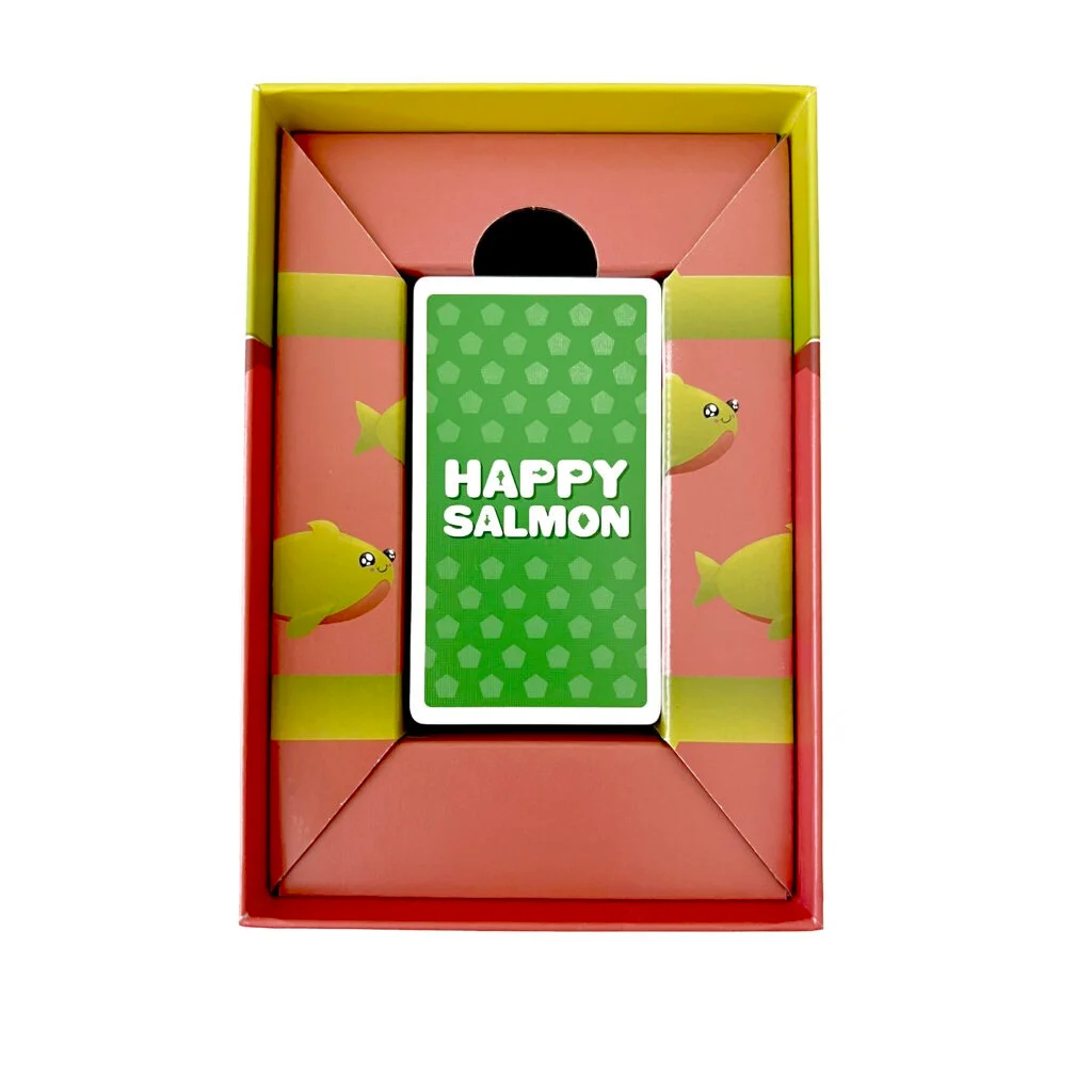 Eine 'Happy Salmon'-Kartenspielbox. Die Box hat ein rotes Innenleben mit gelben Fischen. Eine grüne Karte mit dem Titel befindet sich im Inneren.
