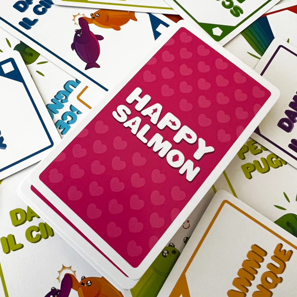 Ein Kartenspiel zeigt 'Happy Salmon' in weißer Schrift auf einer rosa Karte mit Herzmustern. Andere Karten mit Fischillustrationen sind sichtbar.