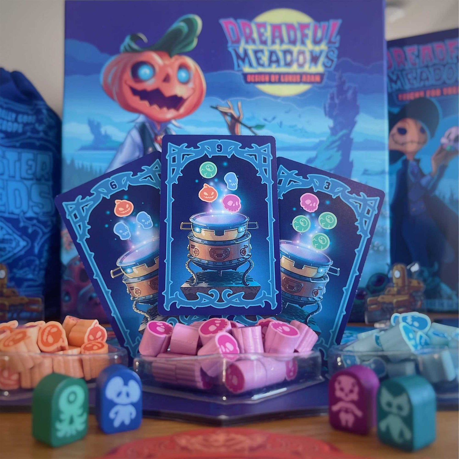 Ein Dreadful Meadows Brettspiel-Setup. Karten mit Kesselkunst, Kürbiskopf-Charakter auf der Box und Spielsteine in Behältern.
