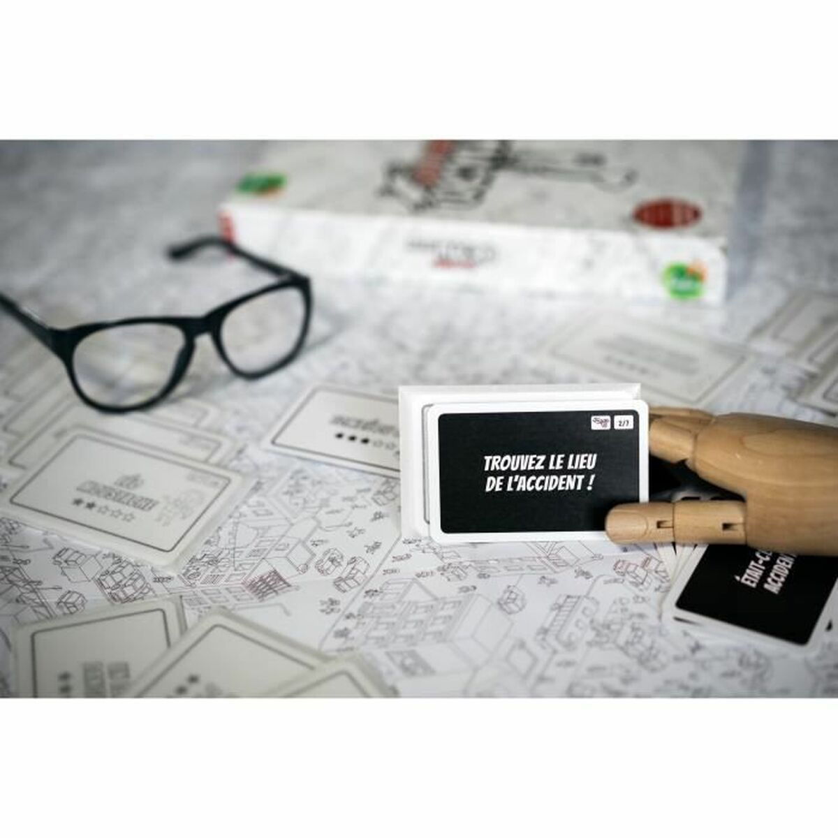 Ein Spiel mit Karten, Brille und Holzhand. Eine Karte zeigt 'Trouvez le lieu de l'accident!' auf einer Karte.