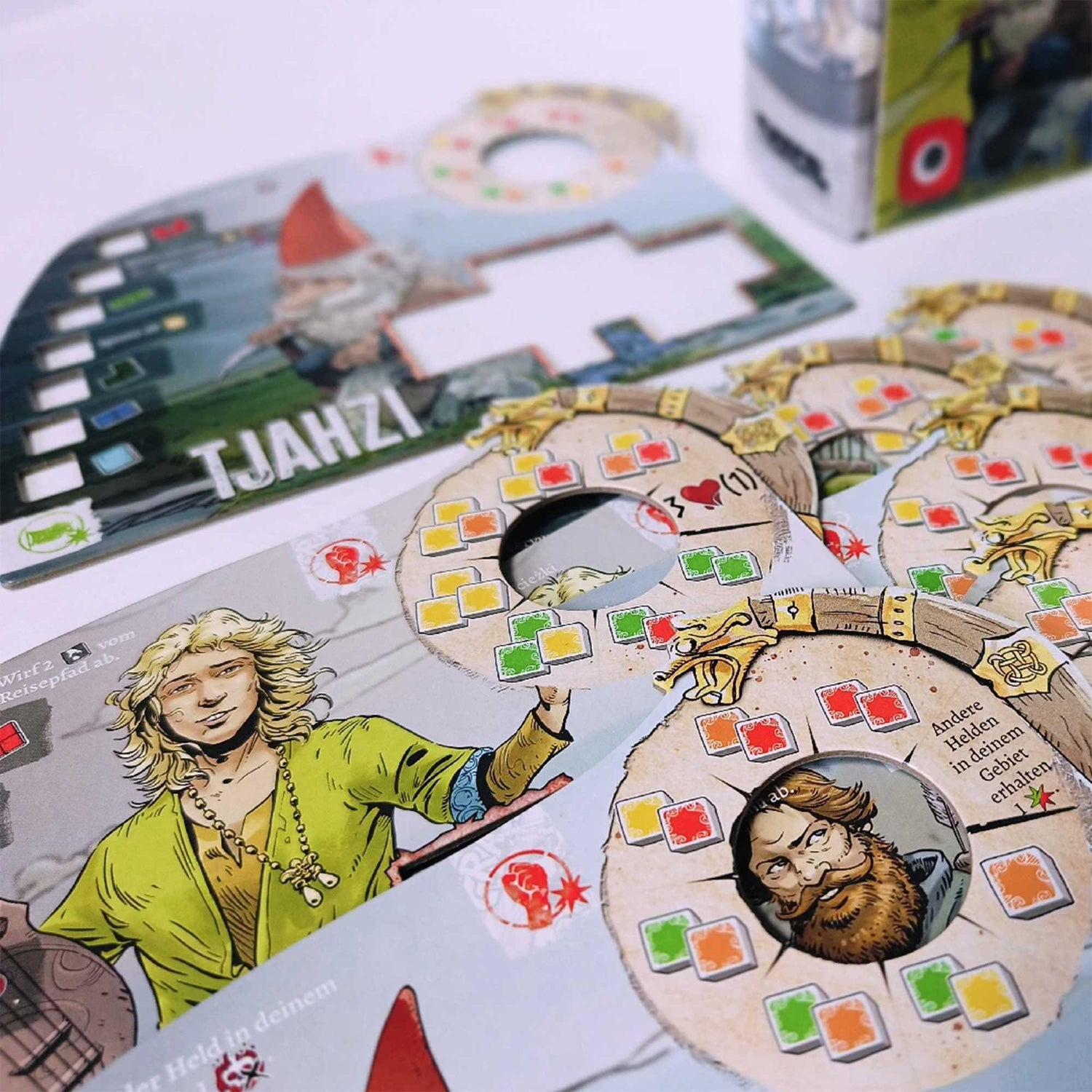 Brettspielkomponenten werden angezeigt. Karten mit Illustrationen von Charakteren, runde Token und eine Spielbox sind sichtbar.