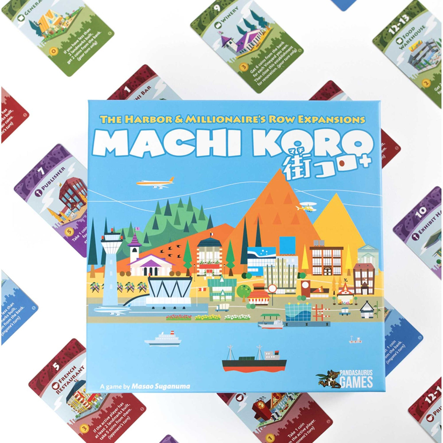 Die Machi Koro Brettspielbox. Gebäude, ein Boot und Karten umgeben die Box. Der Spieletitel ist auf Englisch und Japanisch.