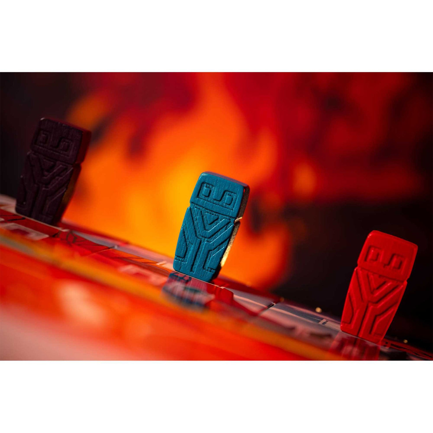Drei bunte Spielsteine auf einem Brett. Die Steine sind blau, rot und schwarz, mit einem feurigen orangefarbenen Hintergrund.