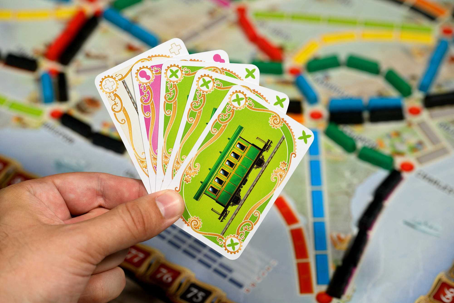 Hand hält Spielkarten mit Zugbildern. Der Hintergrund ist ein Brettspiel mit farbigen Pfaden und Zahlen.