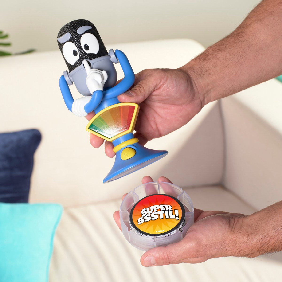 Een persoon houdt een spel vast met een microfoonvormig personage. De spelbasis zegt 'SUPER SSSSTIL!'. Op de achtergrond is een bank.