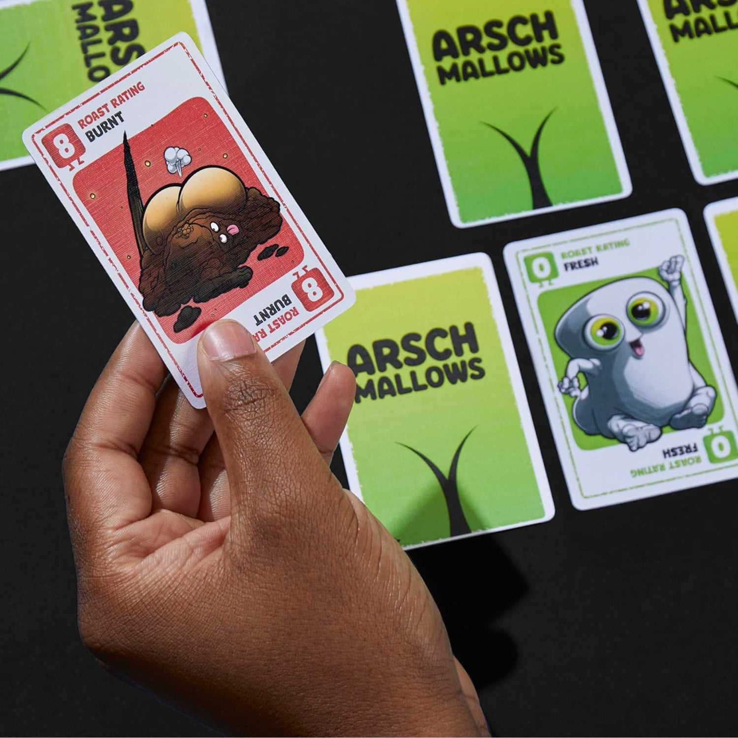 Eine Hand hält eine Spielkarte mit einer Illustration eines verbrannten Marshmallows. Andere Spielkarten sind sichtbar, und der Hintergrund ist schwarz.