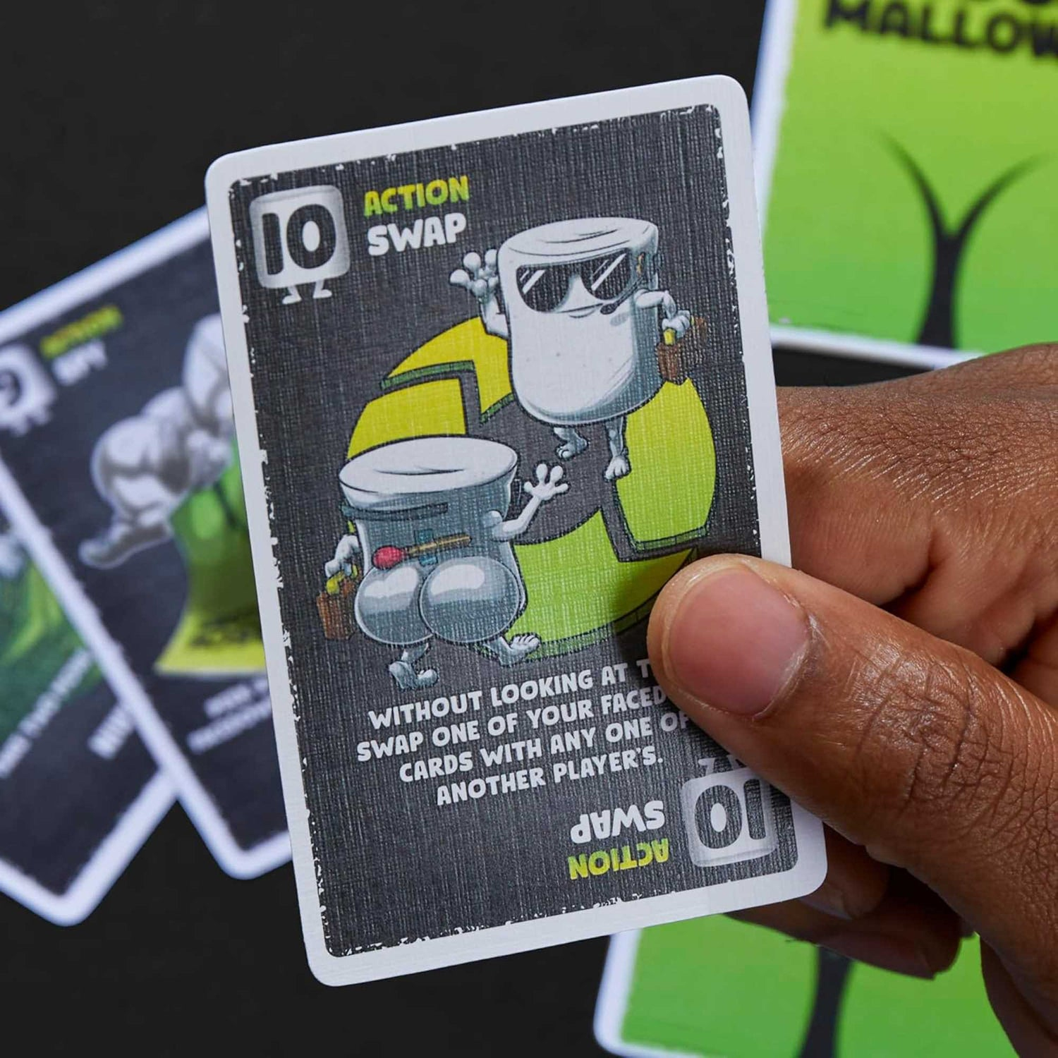 Eine Spielkartenkarte, die von einer Hand gehalten wird. Sie zeigt zwei Zeichentrickfiguren aus Marshmallows und lautet 'Swap'. Die Karte ist schwarz und weiß.