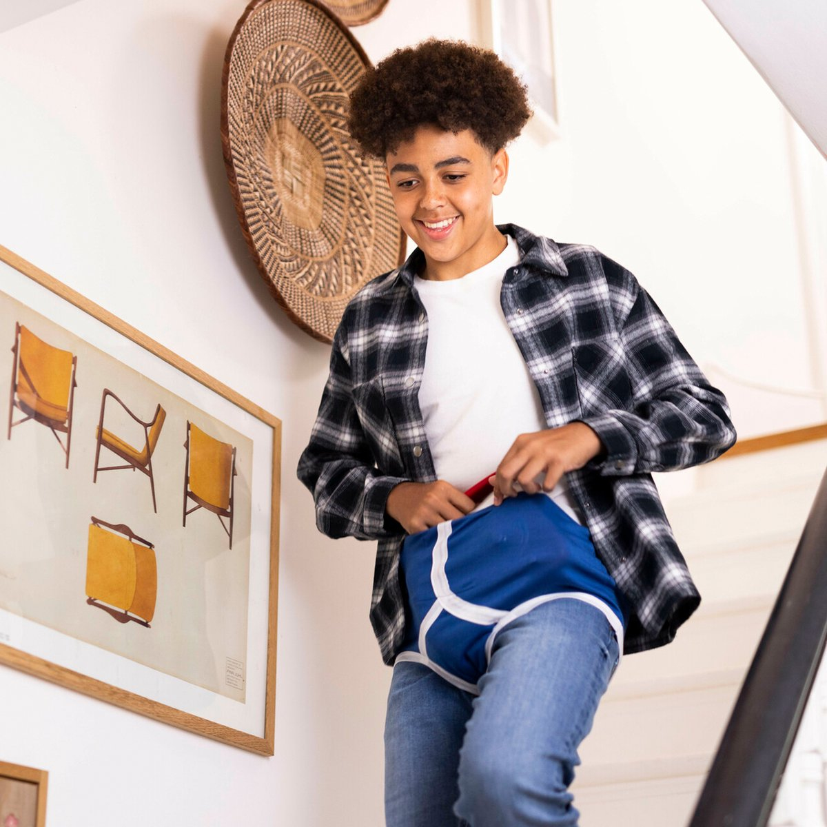 Een lachende tiener in jeans en een geruit overhemd trekt blauw ondergoed met witte randen omhoog. Kunstwerken en geweven decoratie sieren de muur.