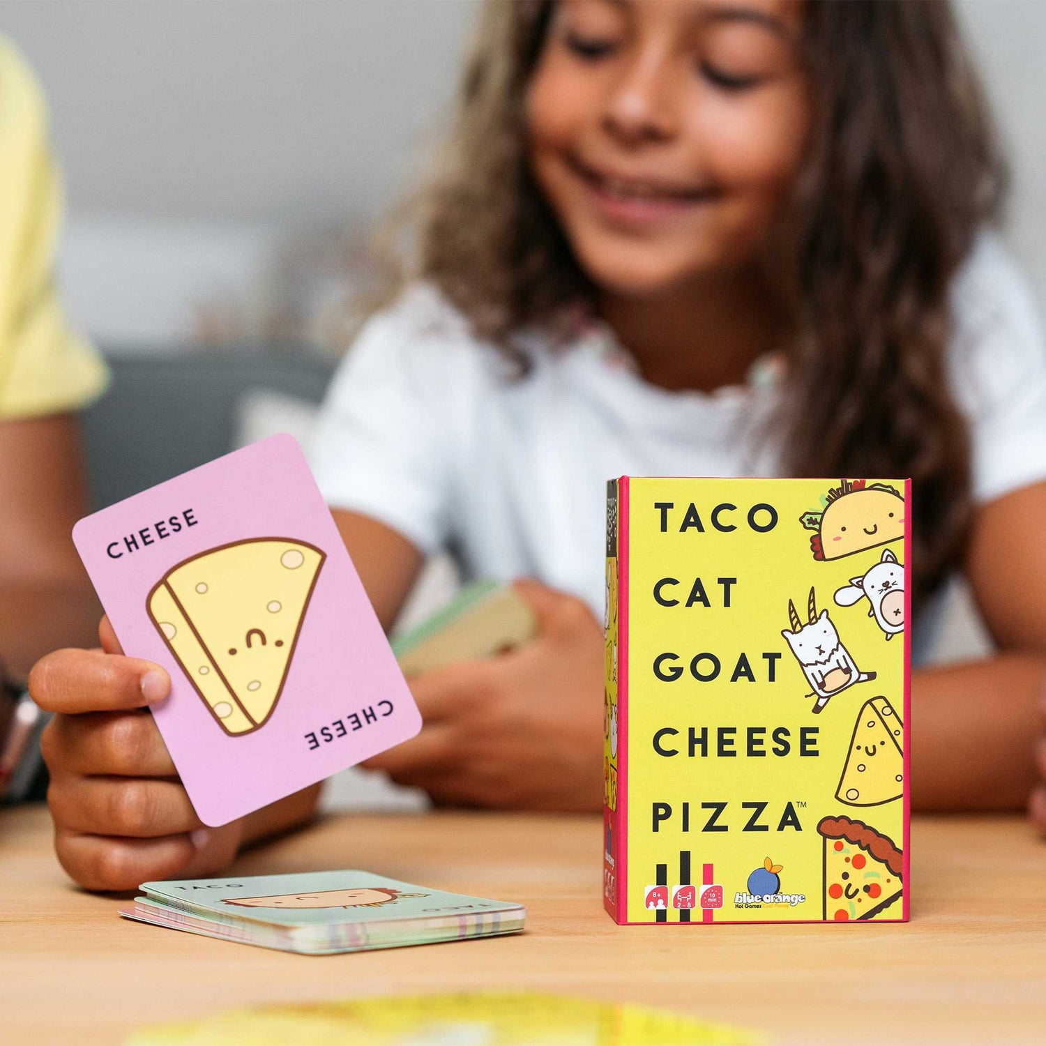 Ein lächelndes Mädchen hält eine rosafarbene Karte mit einem Käsebild. Eine Spielbox von Taco Cat Goat Cheese Pizza liegt auf dem Tisch.