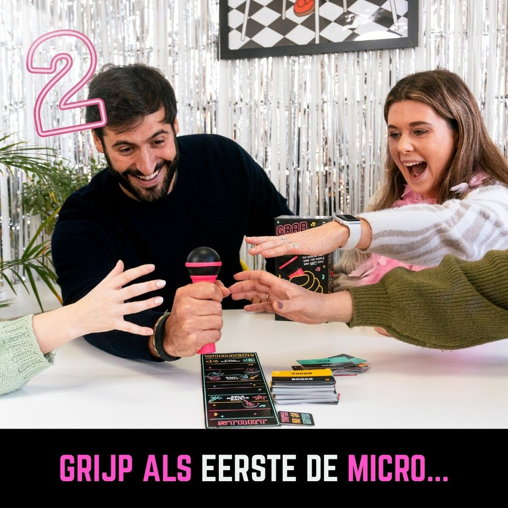 Vier mensen spelen een spel. Een man houdt een roze microfoon vast. Kaarten en een spelbord liggen op tafel. De vrouw schreeuwt van opwinding.