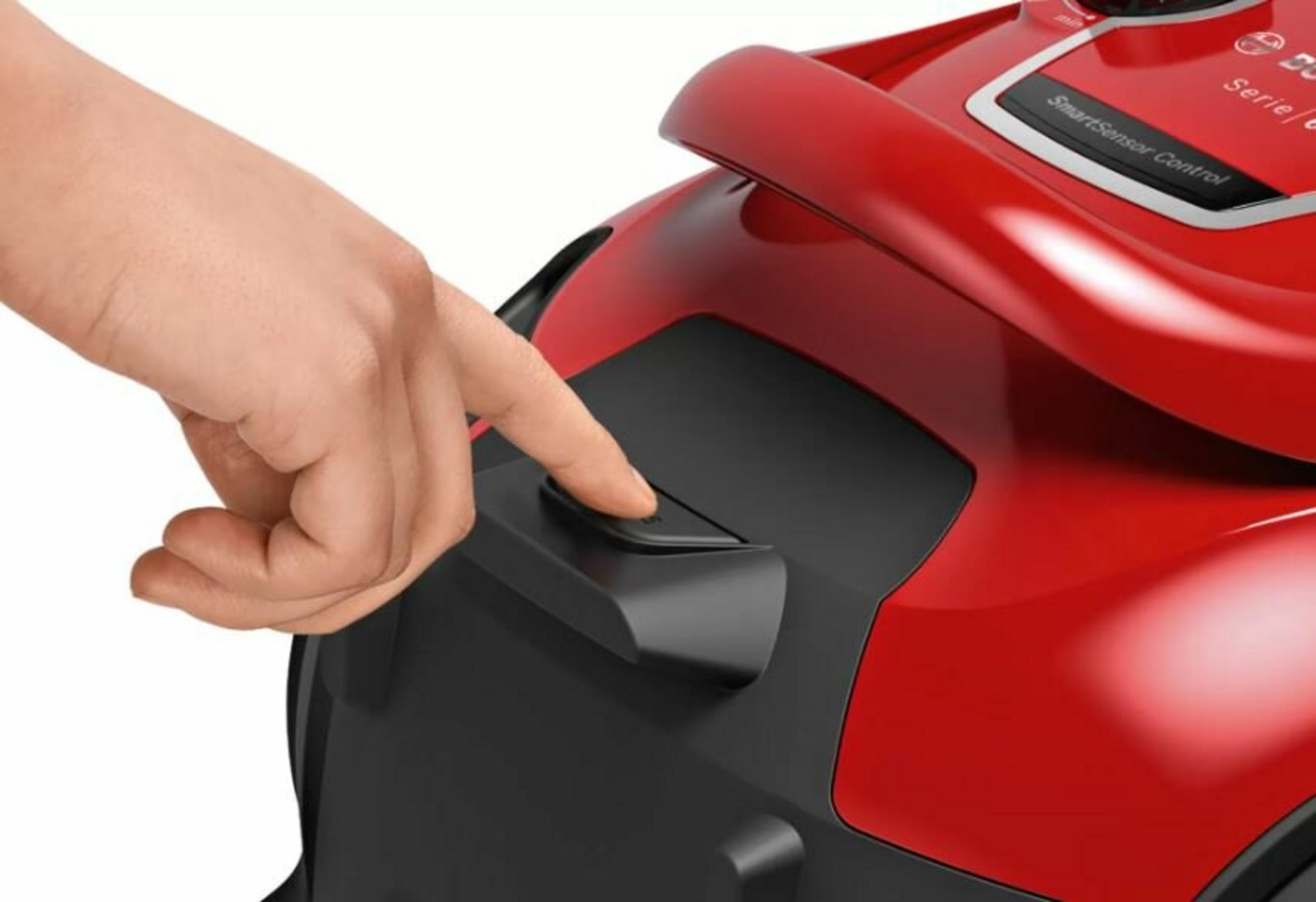 A hand presses a button on a red vacuum cleaner. The button is black. Part of the vacuum is black.