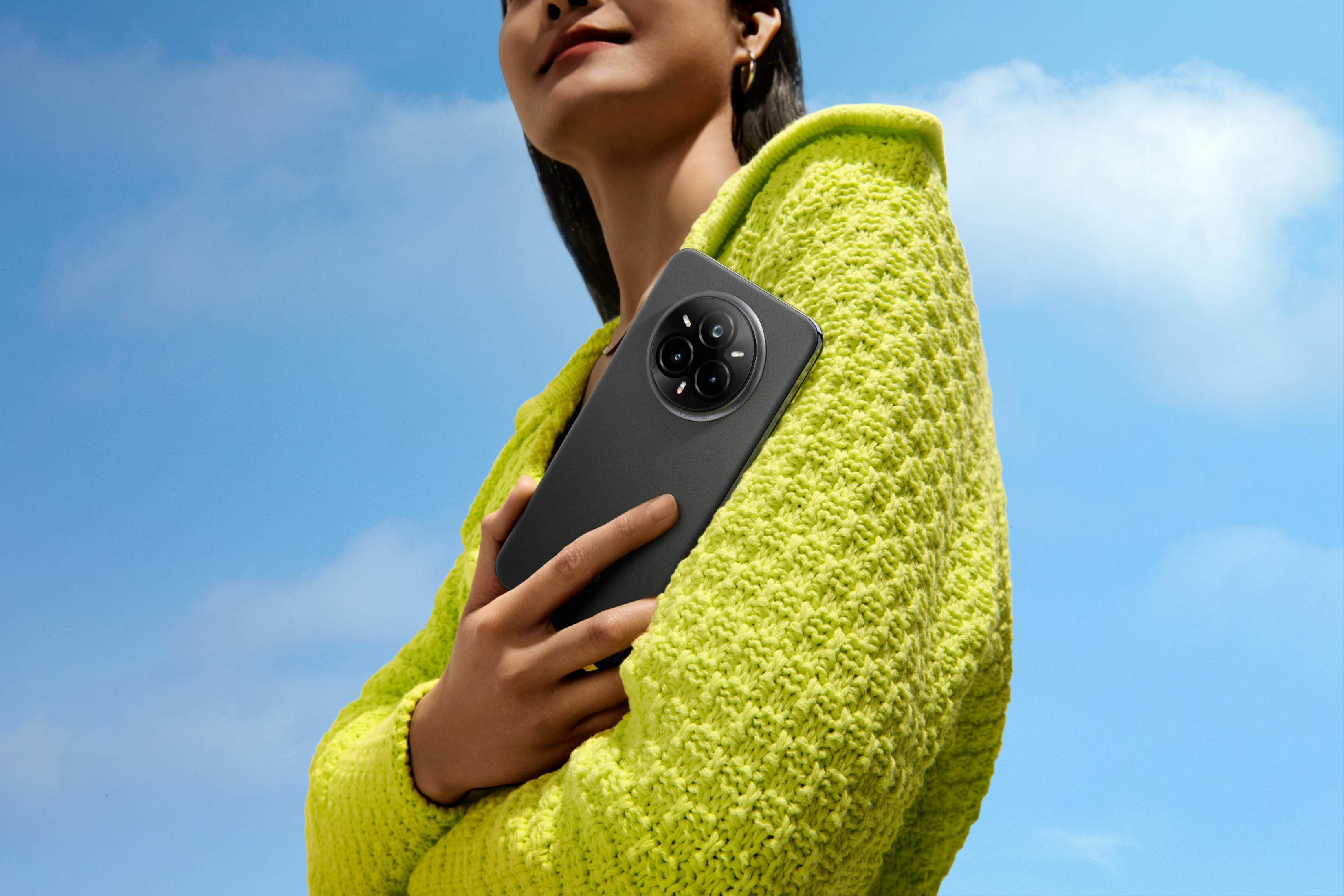 Osoba trzymająca czarny smartfon z wieloma aparatami, ubrana w zielony sweter. Tło niebieskiego nieba.