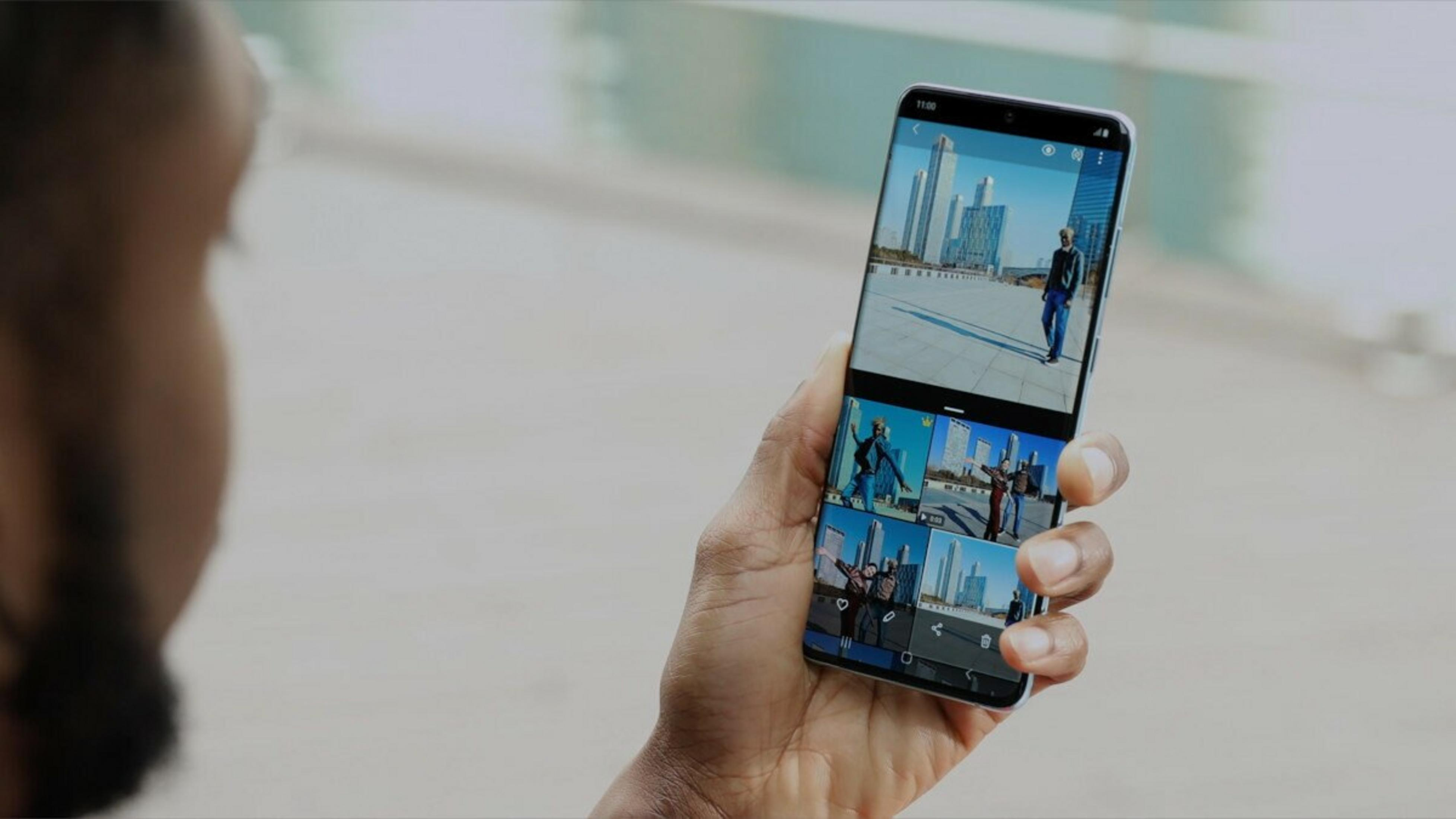 Una mano tiene un telefono che mostra foto di una persona di fronte a degli edifici. Lo sfondo è sfocato, con la spalla di una persona visibile.