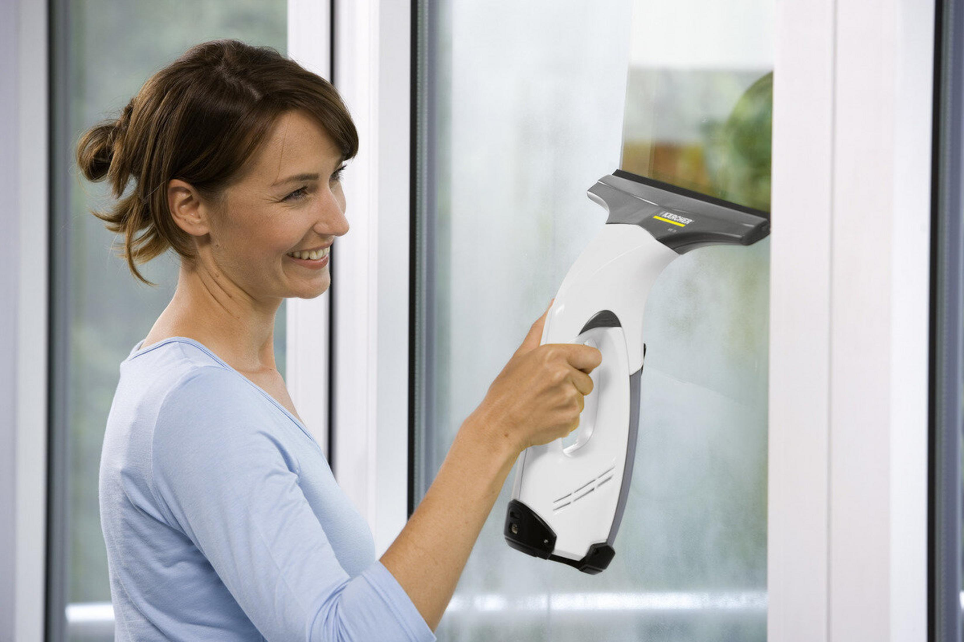 Kobieta czyści okno biało-szarą myjką do okien. Uśmiecha się. Tło to okno i biała rama.