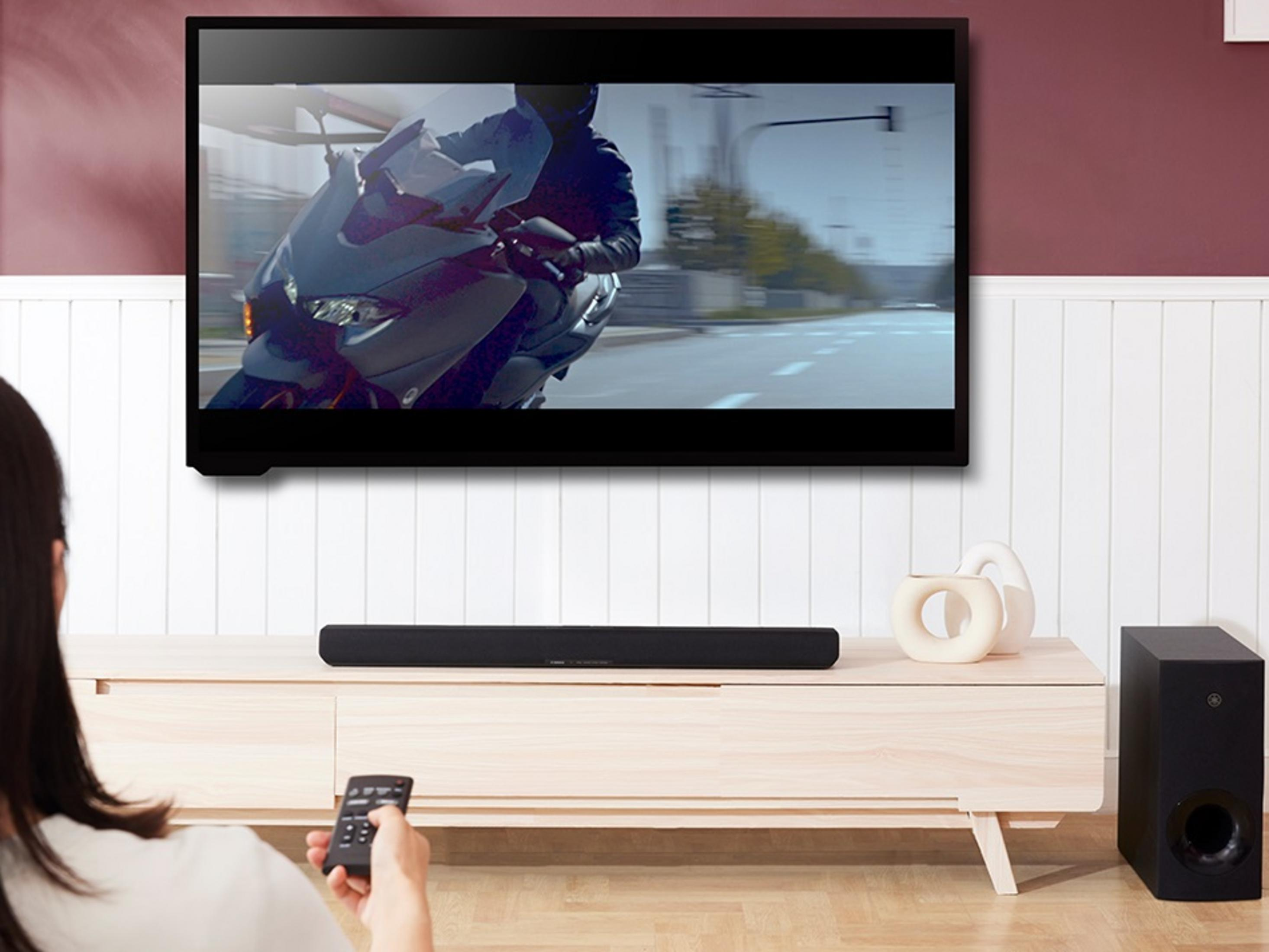 A woman using a remote. TV shows a motorcycle. Soundbar, subwoofer, and decor are on a wooden shelf.