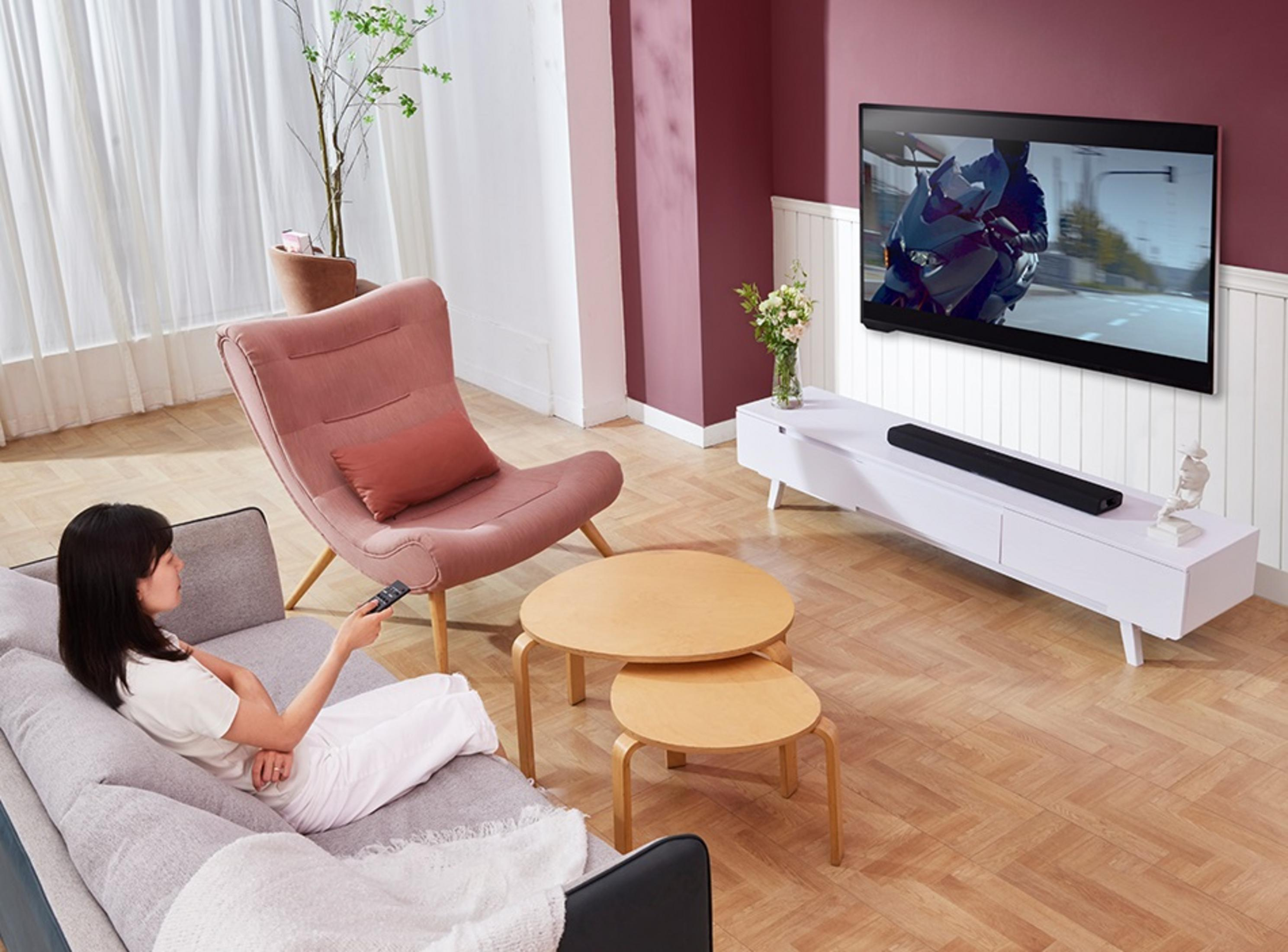 Una donna sul divano guarda la TV. La stanza ha una sedia rosa, tavoli di legno e un mobile TV bianco.