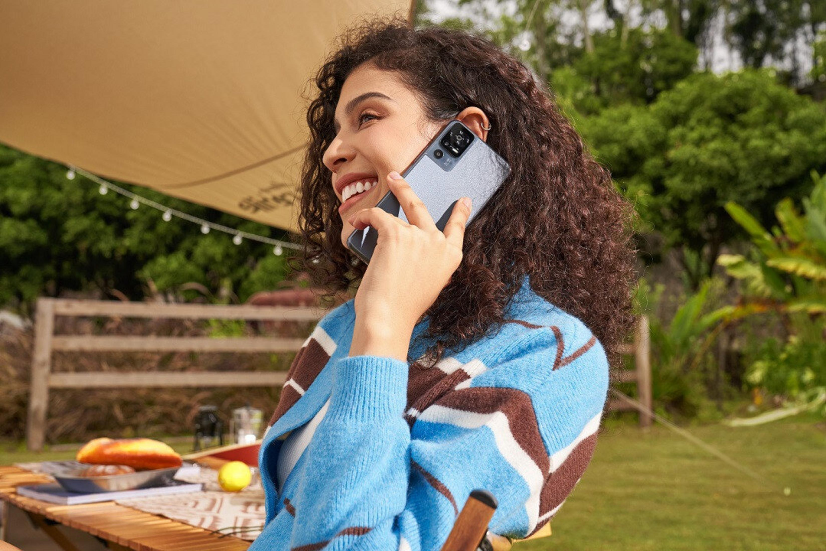 Una mujer sonríe mientras habla por teléfono. Lleva un suéter azul y marrón, con cabello rizado. Un teléfono en su oído.