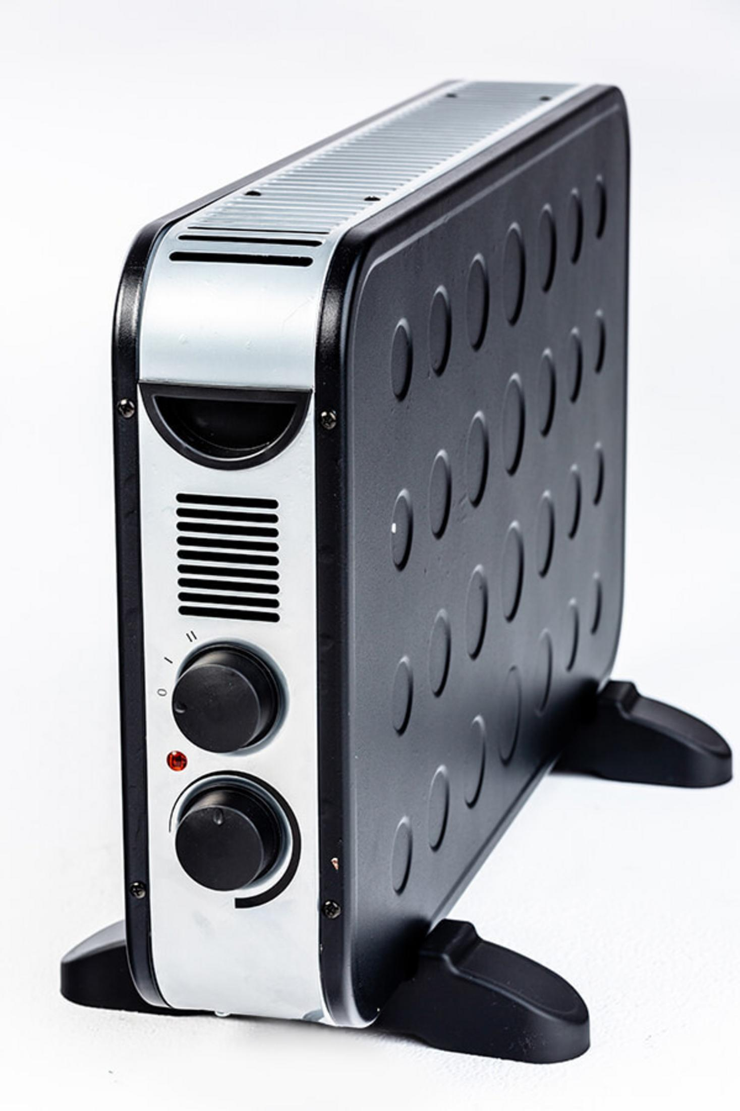 Czarno-srebrny grzejnik elektryczny. Ma dwa pokrętła, czerwone światło i owalne otwory wentylacyjne. Stoi na dwóch czarnych nóżkach.