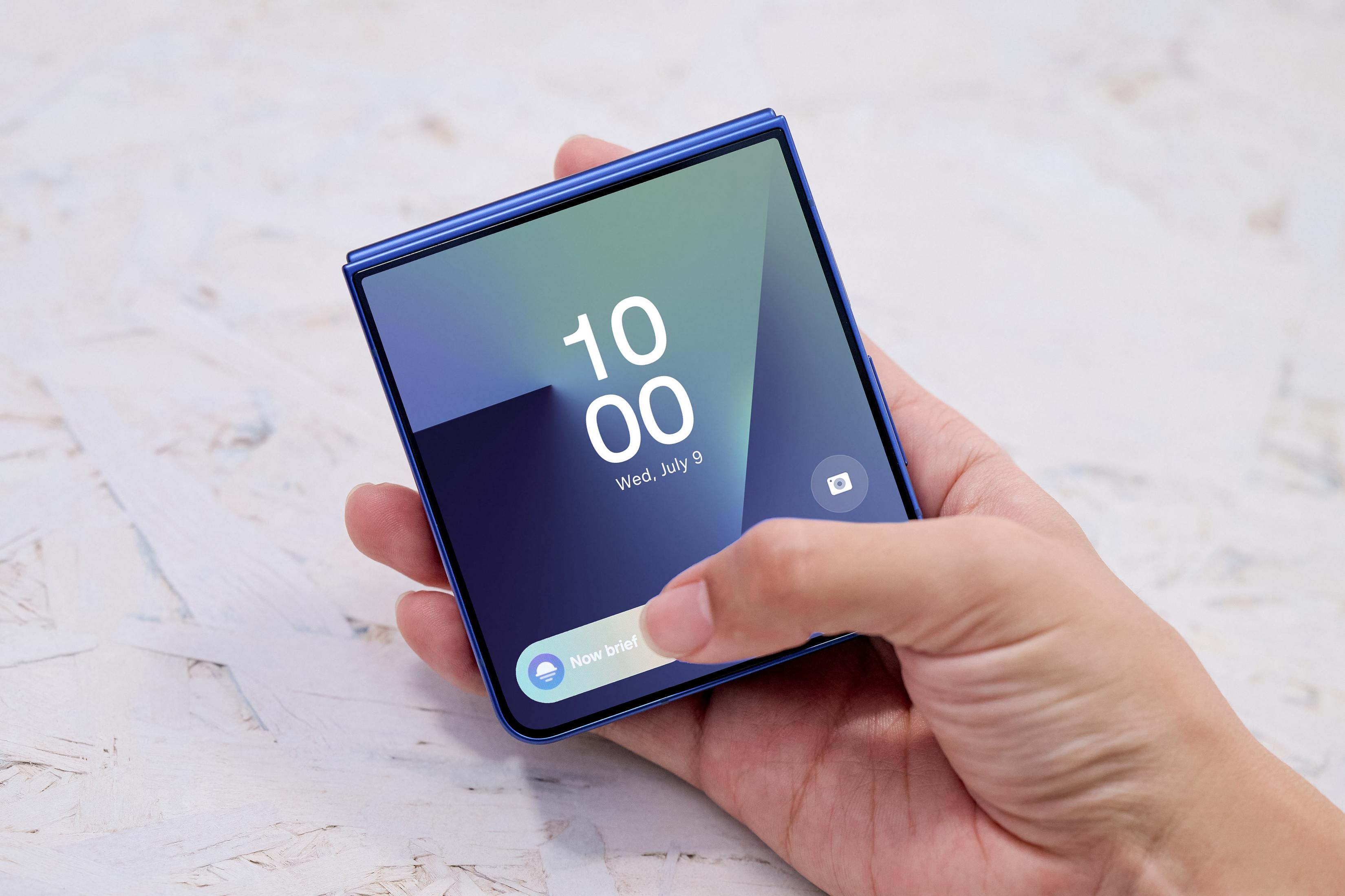 Eine Hand hält ein blaues Smartphone. Der Bildschirm zeigt Uhrzeit, Datum und die Schaltfläche 'Now brief'.
