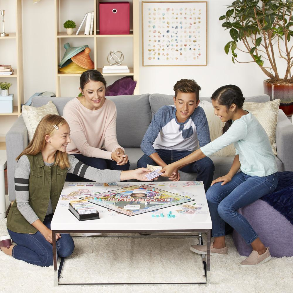 Vier Personen spielen Monopoly. Ein Brettspiel steht auf einem weißen Tisch. Sie sitzen auf einem Sofa und dem Boden.