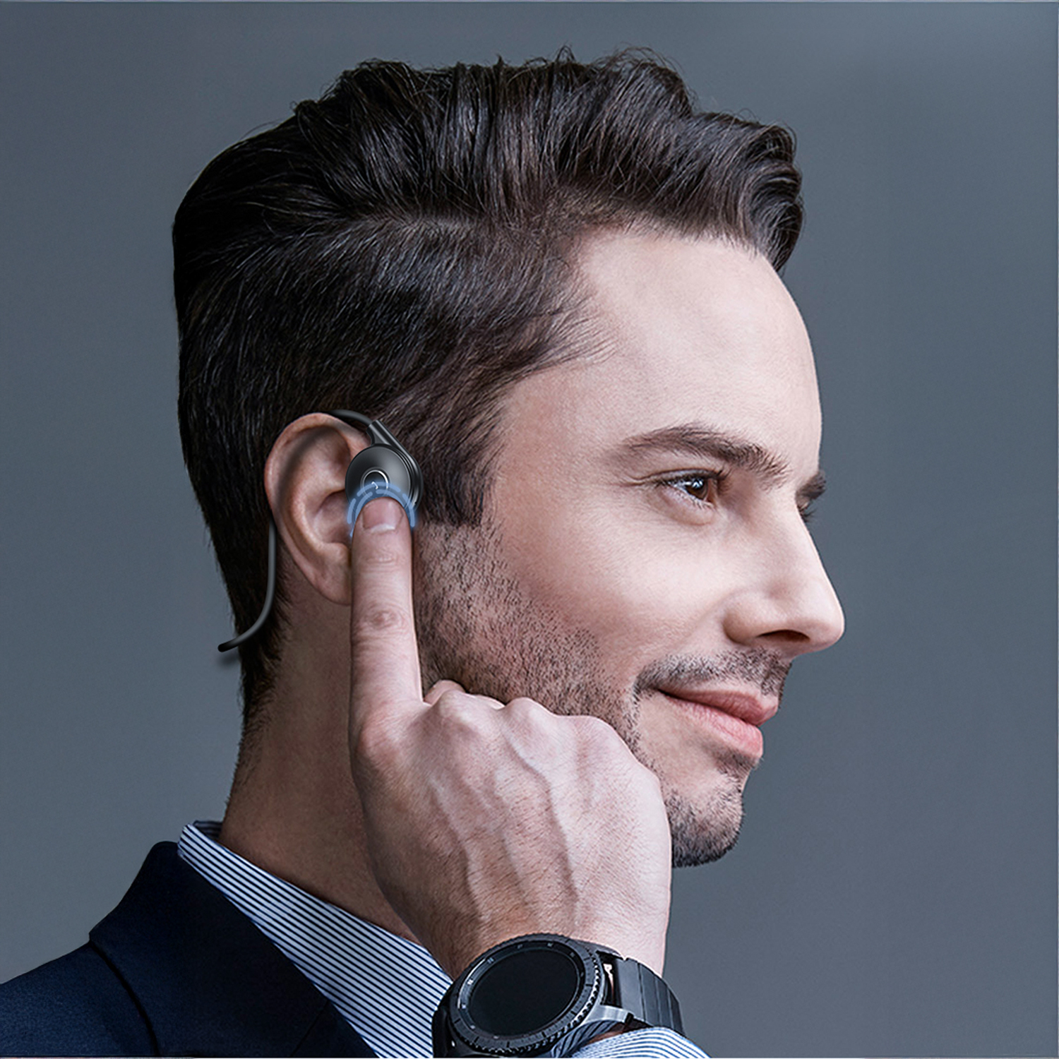 Un uomo con un completo e uno smartwatch regola un auricolare. L'uomo ha i capelli castani e sorride. Lo sfondo è grigio.