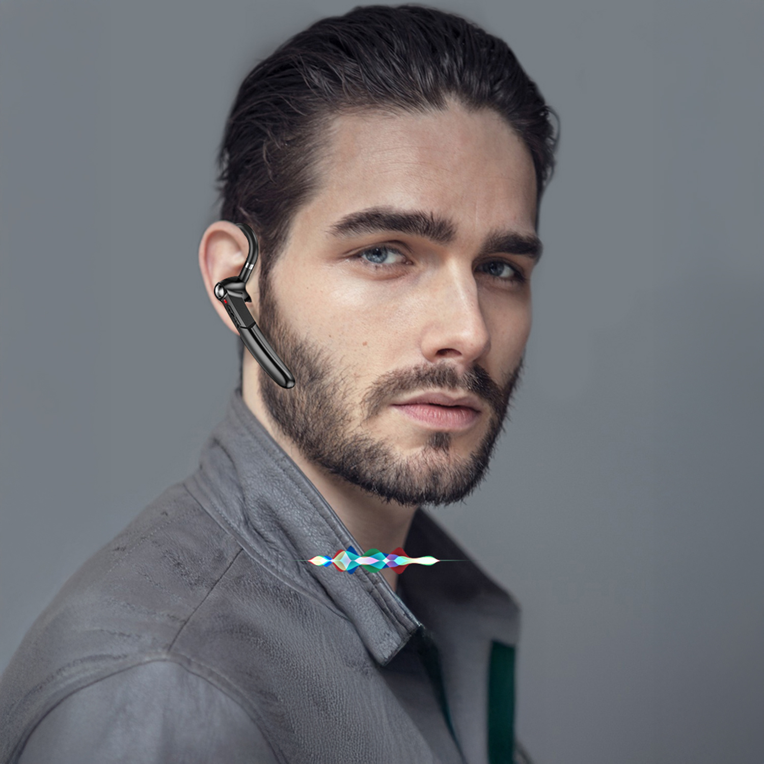 Un uomo con la barba indossa un auricolare. Indossa una giacca di pelle grigia. Lo sfondo è grigio.
