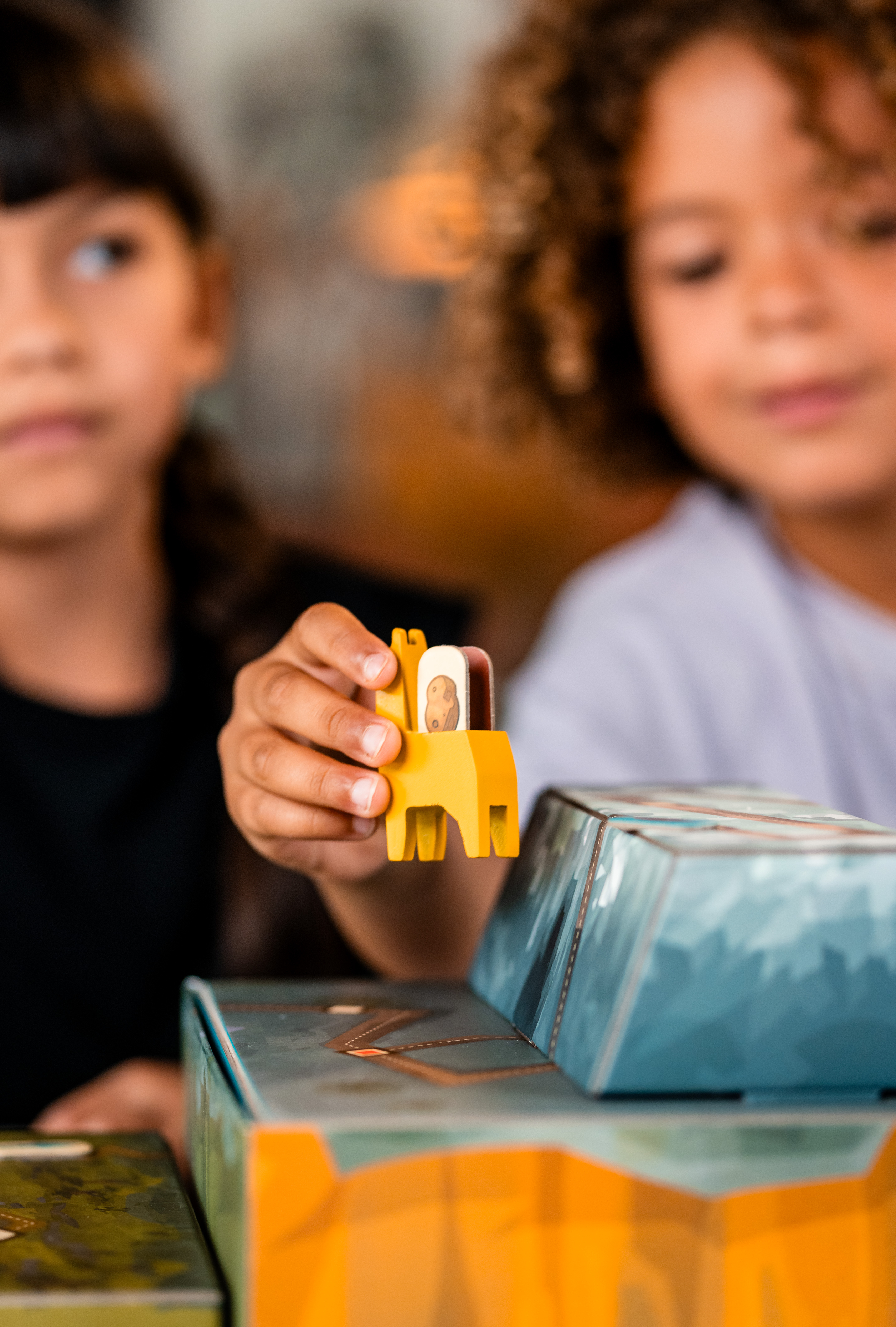 Twee kinderen spelen een spel. Eén houdt een geel houten paard vast. Blauwe en groene dozen op de voorgrond.