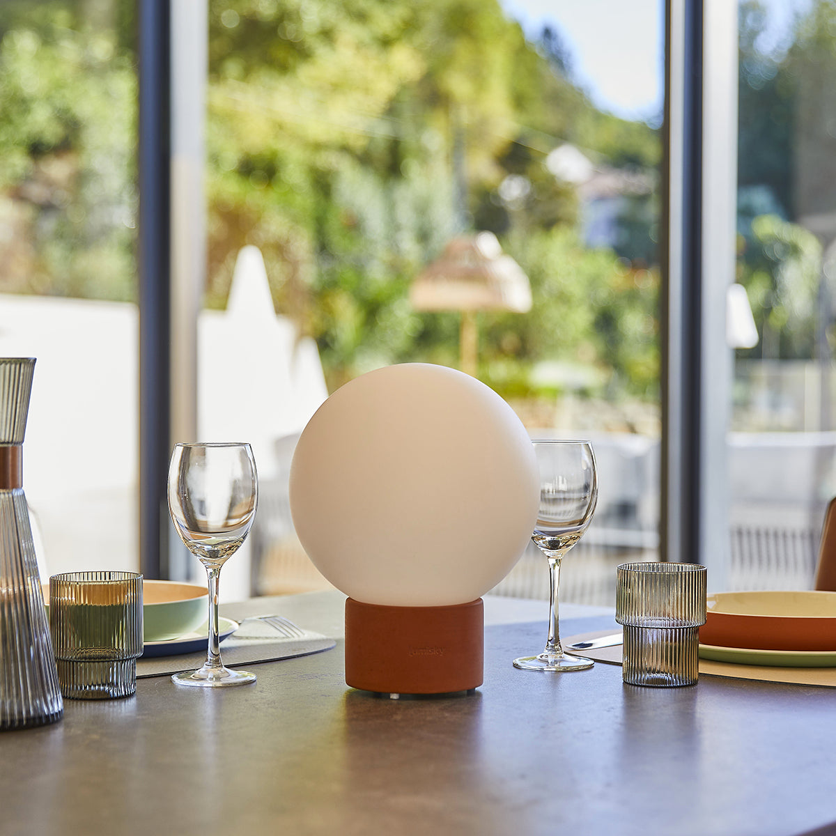 Ein Tisch ist mit Gläsern, Tellern und einer Lampe gedeckt. Ein großes Fenster zeigt eine verschwommene Ansicht von grünen Bäumen im Hintergrund.