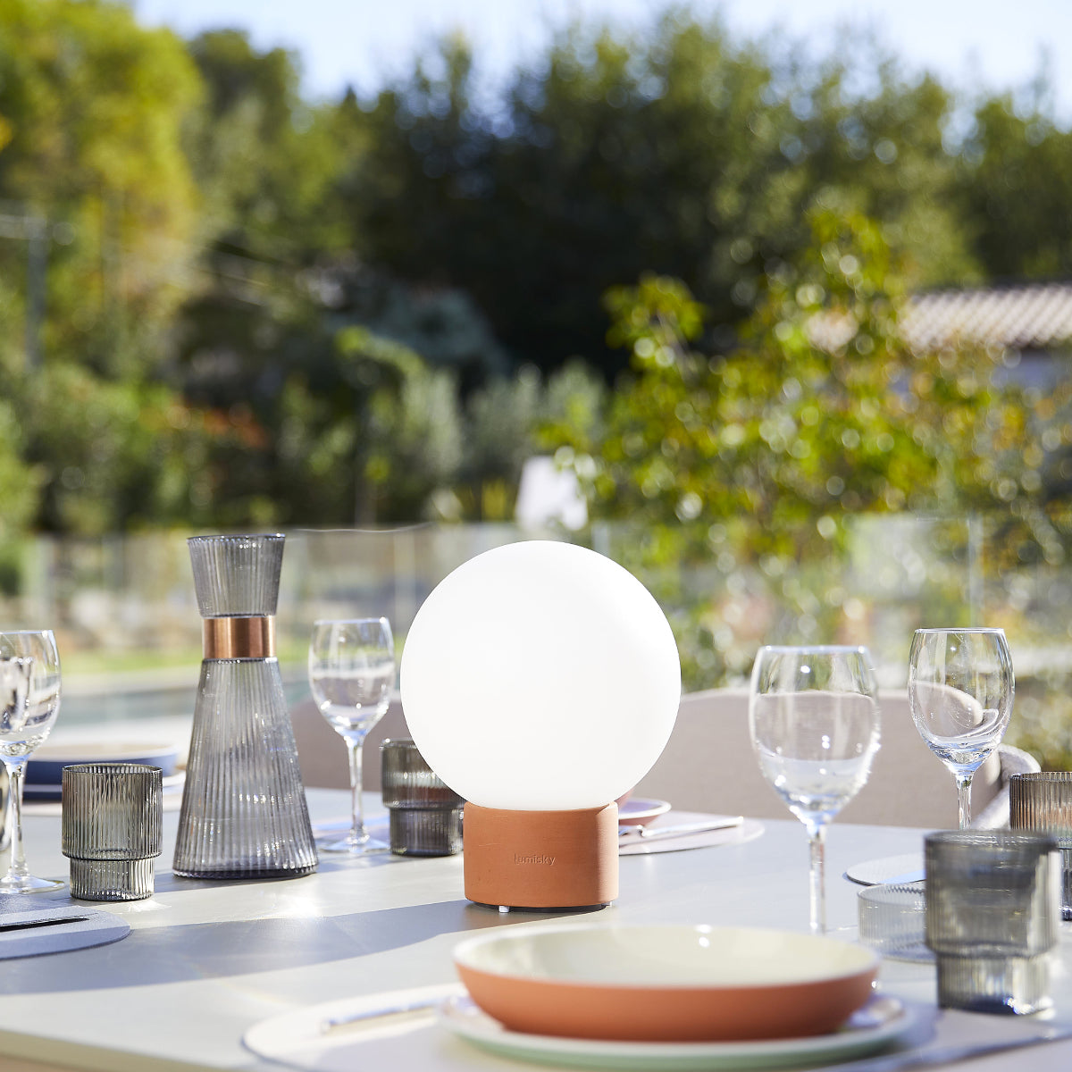Gedeckter Tisch im Freien mit Globuslampe, Gläsern und Tellern. Im Hintergrund Bäume und ein Haus, sonniger Tag.