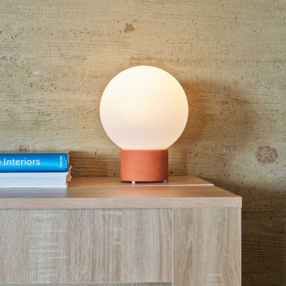 Eine runde, weiße Lampe steht auf einem Holztisch. Links ist ein Buch mit dem Titel 'e Interiors'. Der Hintergrund ist eine strukturierte Wand.