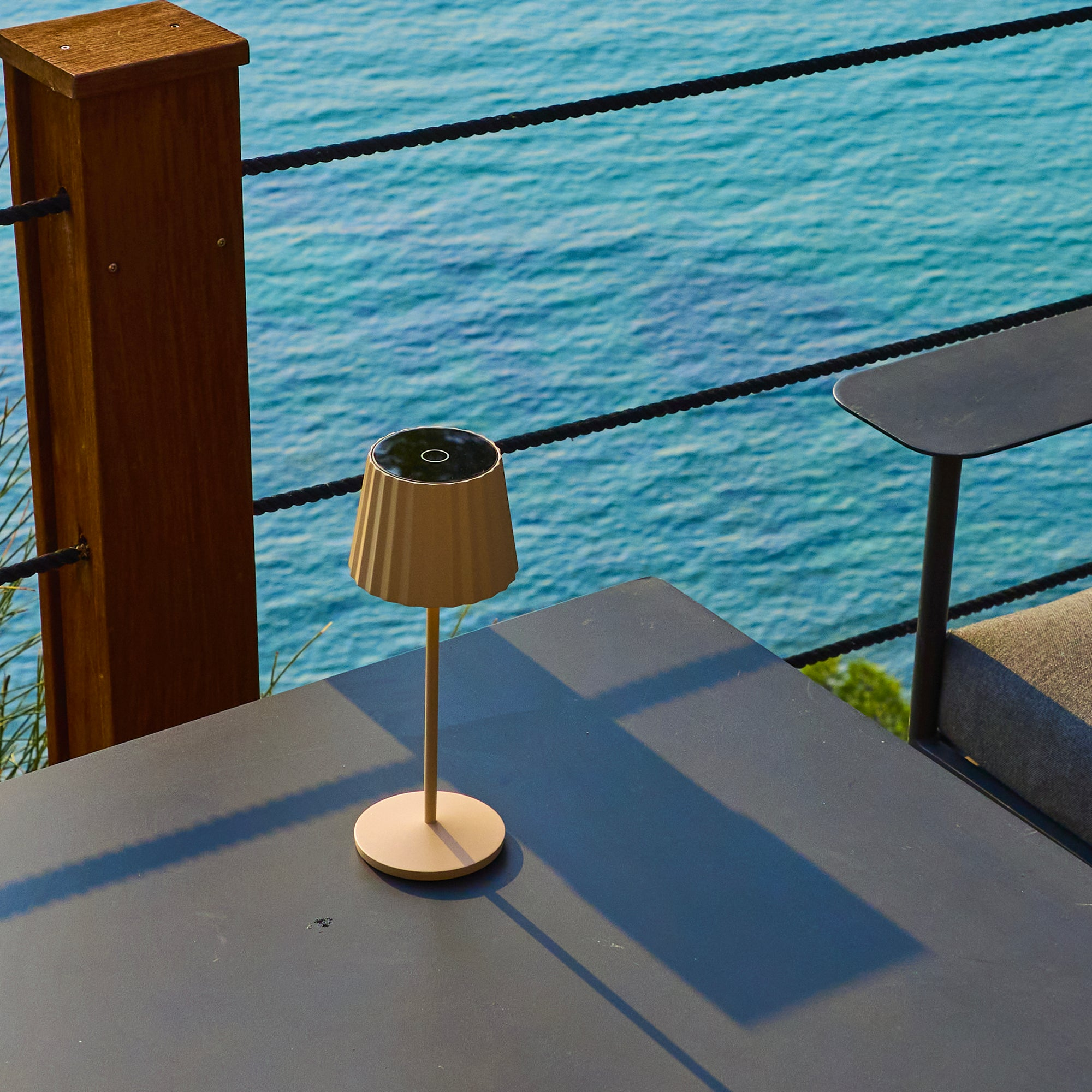 Eine beige Lampe auf einem dunklen Tisch, Meer im Hintergrund. Schwarzer Seilzaun und Holzpfosten.