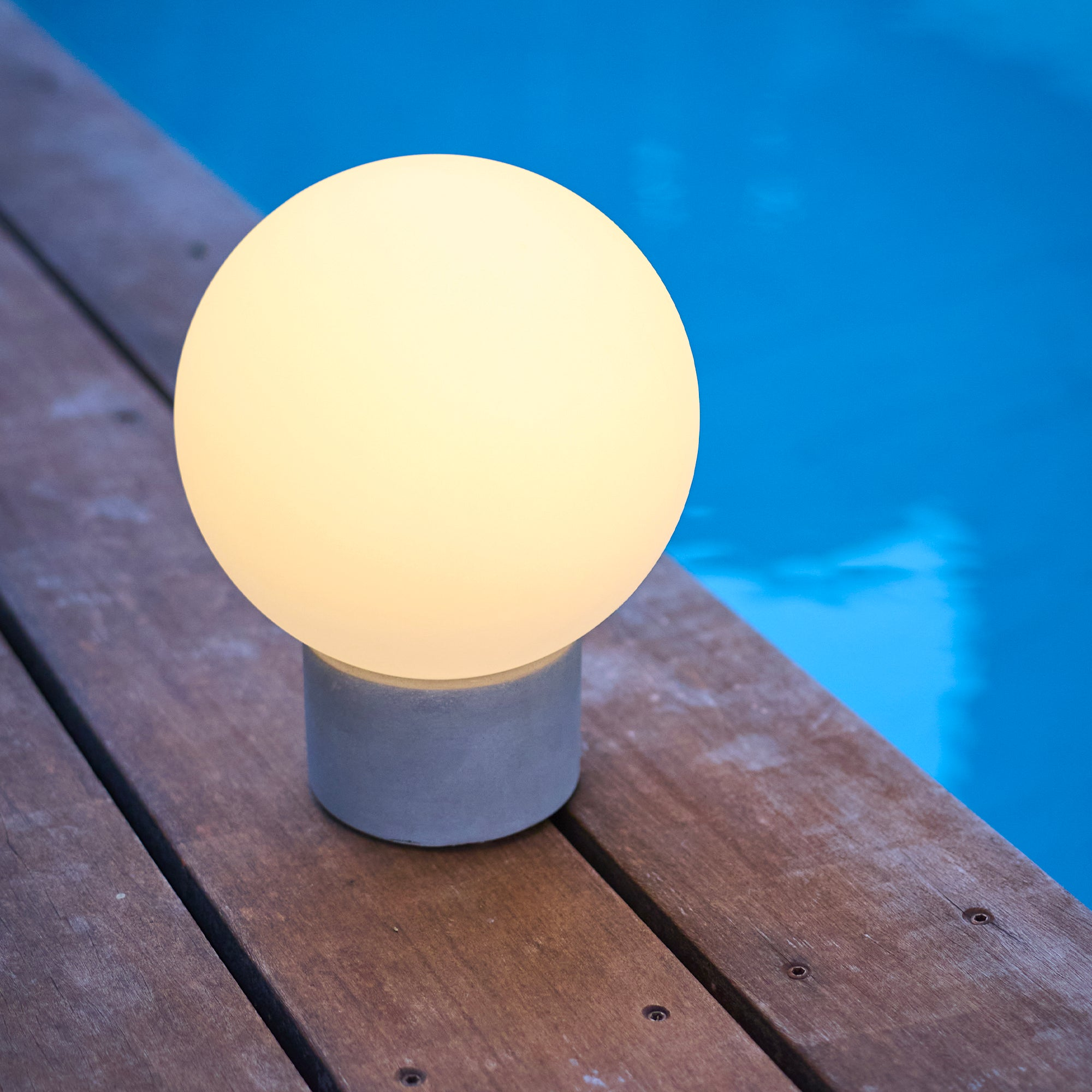 Eine leuchtende Kugellampe auf einem Holzdeck, mit einem Pool im Hintergrund. Die Lampe ist weiß und rund, der Pool ist blau.