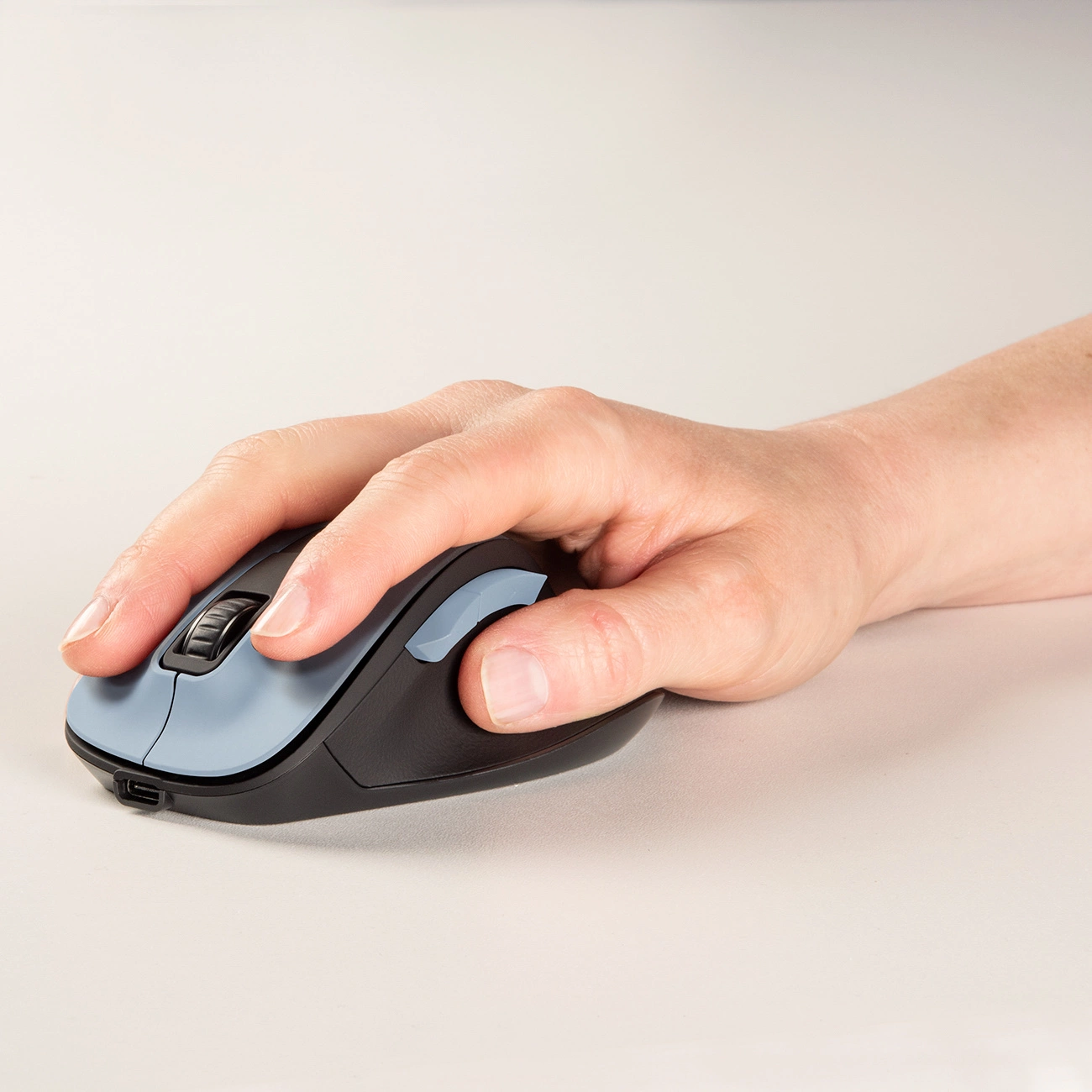 Una mano poggia su un mouse per computer. Il mouse è nero e blu. Lo sfondo è una superficie liscia, bianco sporco.