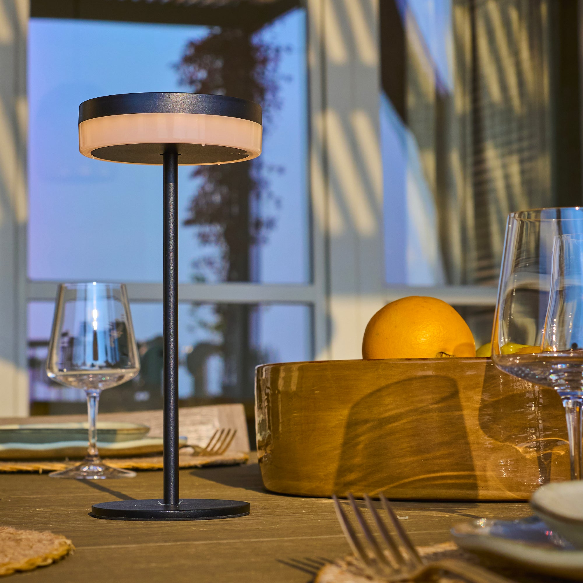Eine Tischlampe mit schwarzem Sockel und rundem Licht. Es gibt eine Holzschale mit einer Orange und Weingläsern.