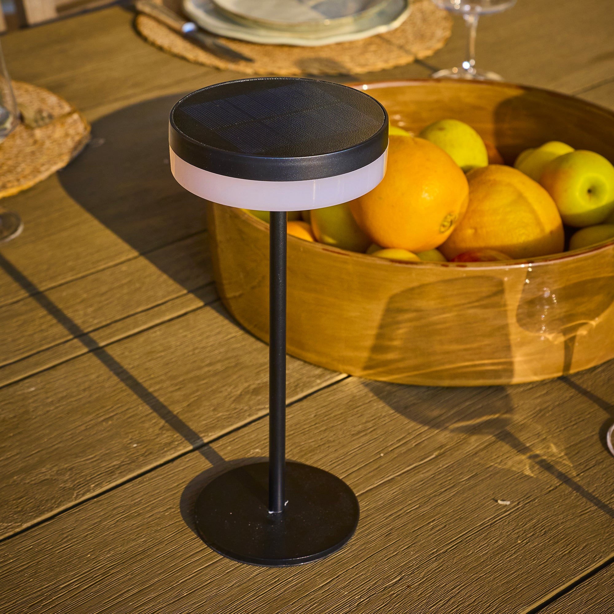 Eine Solarlampe auf einem Holztisch, im Hintergrund eine Obstschale mit Orangen und grünen Äpfeln.