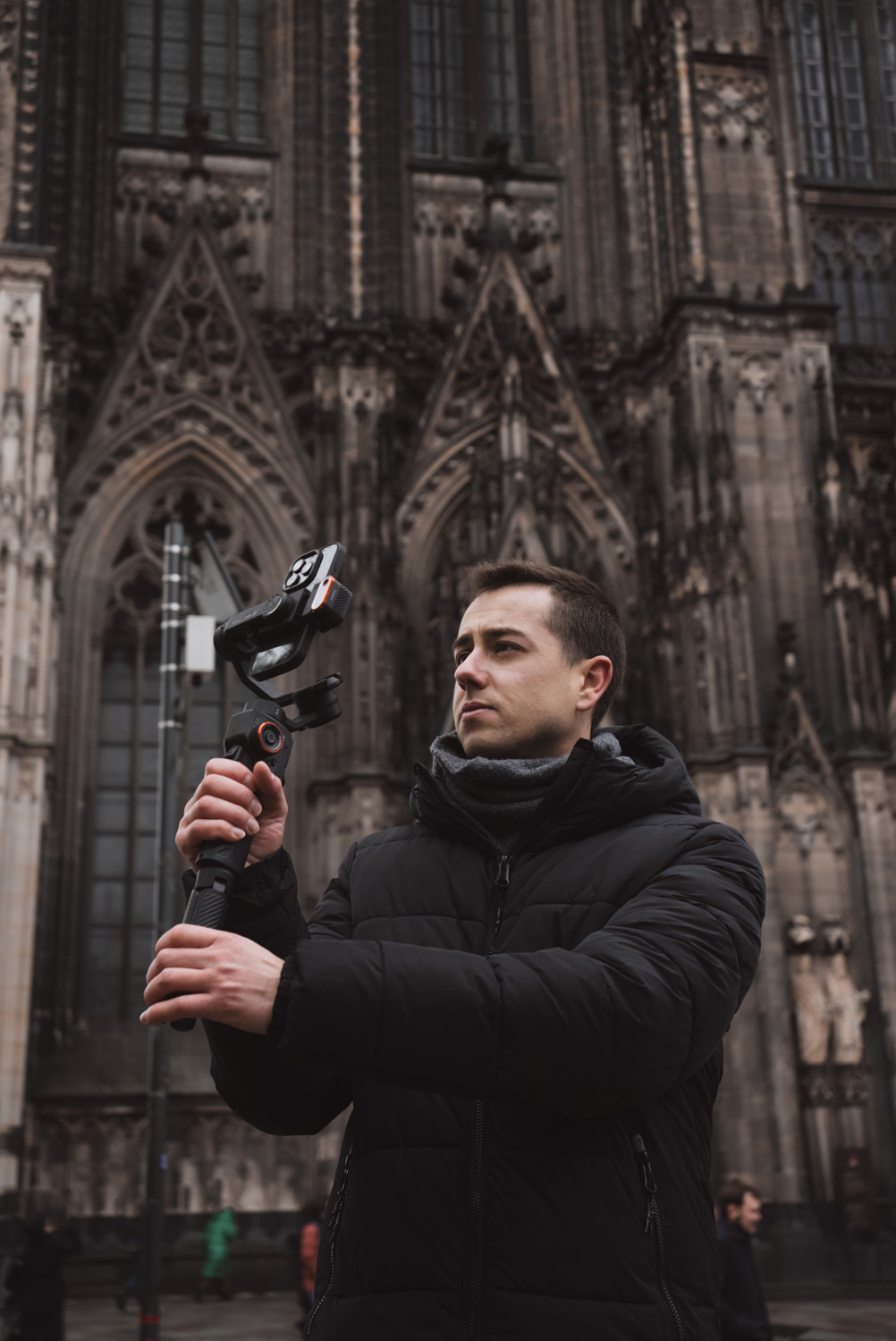 Mężczyzna trzyma stabilizator kamery przed katedrą. Ma na sobie czarną kurtkę i patrzy w górę. Tłem jest katedra.