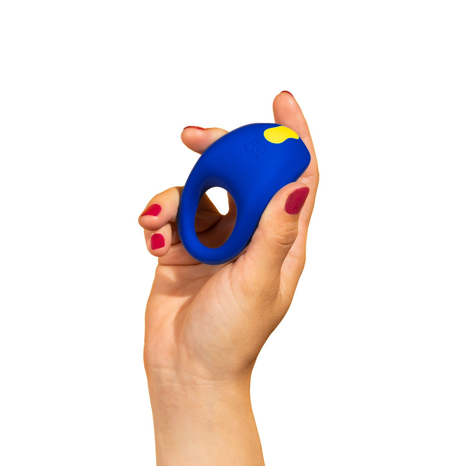 Una mano sostiene un juguete sexual azul en forma de anillo con un botón amarillo. La mujer tiene esmalte de uñas rojo. Fondo blanco.