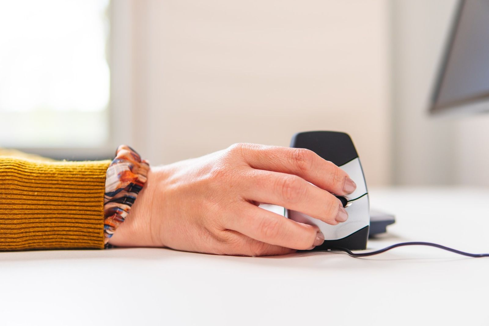 A hand wearing a bracelet is using a computer mouse. The mouse is black and silver, connected by a black wire.