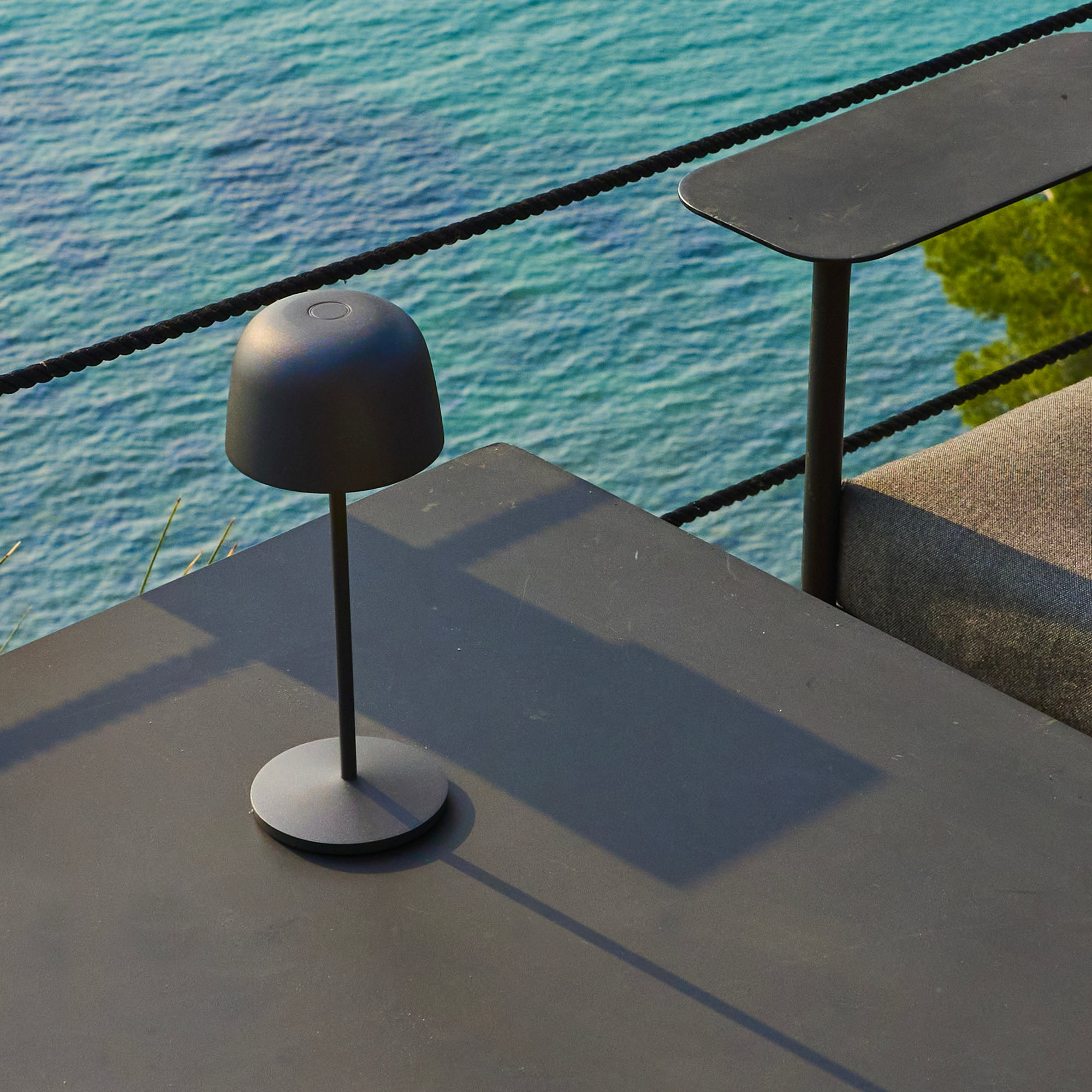 Eine schwarze Lampe steht auf einem dunklen Tisch, im Hintergrund Meerblick. Ein zweiter Tisch und Seil sind sichtbar.