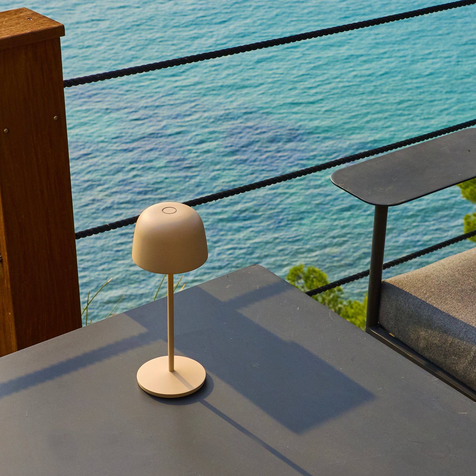 Eine beige Lampe auf einem dunkelgrauen Tisch mit Meerblick im Hintergrund. Rechts ist ein Stuhl.