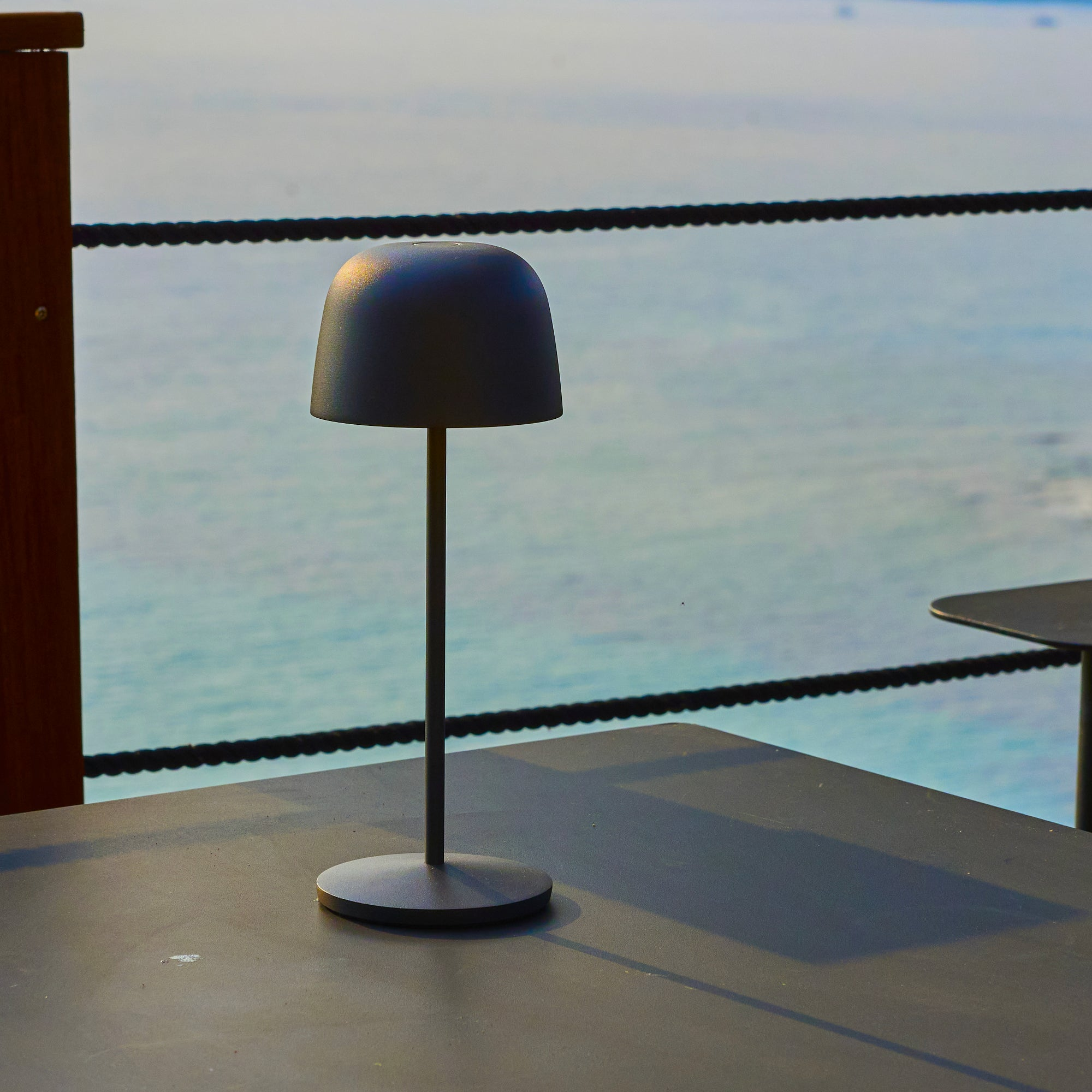 Eine schwarze Lampe auf einem Tisch mit Meerblick im Hintergrund. Ein Seilzaun trennt den Tisch vom Meer.