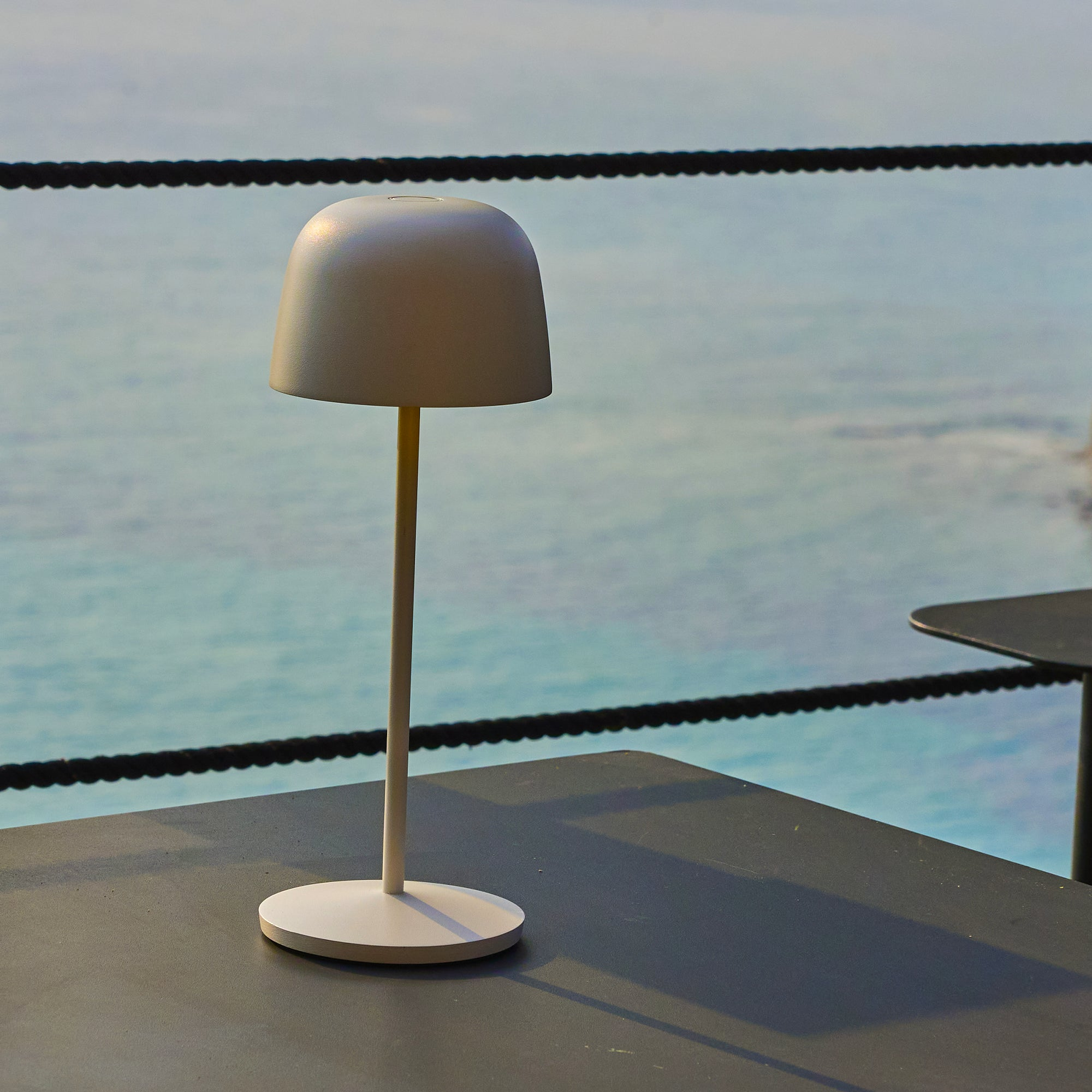 Eine weiße Lampe auf einem dunklen Tisch, mit Meer im Hintergrund. Oben ist ein schwarzes Seil zu sehen.