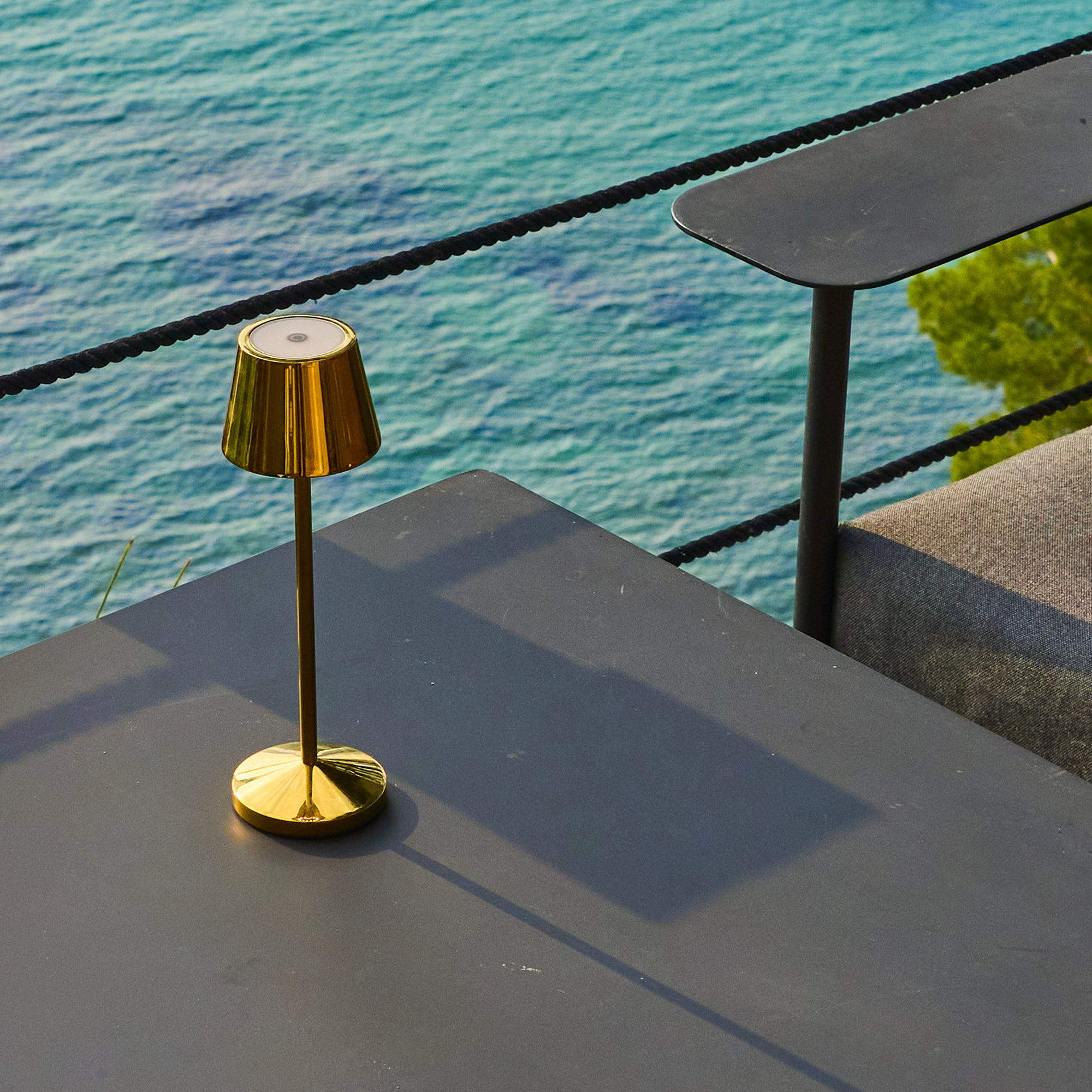 Goldene Lampe auf Tisch mit Meerblick. Schwarzes Seil und Tisch sichtbar. Das Meer ist blau.