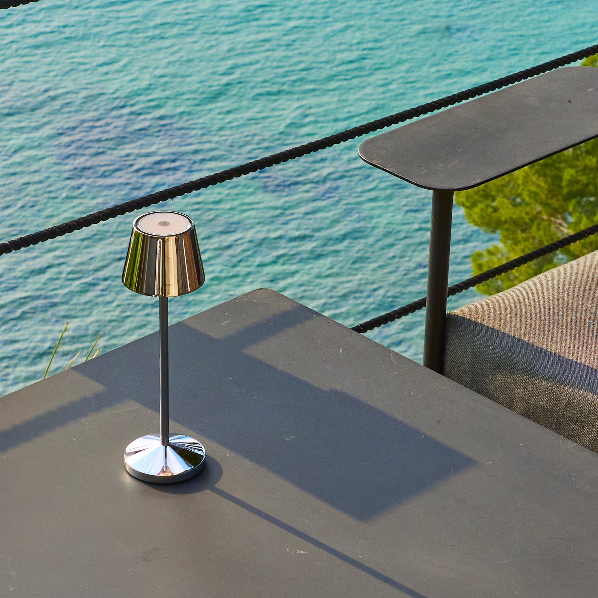 Eine Tischlampe auf einem dunklen Tisch, mit Meer und schwarzem Seilzaun im Hintergrund.