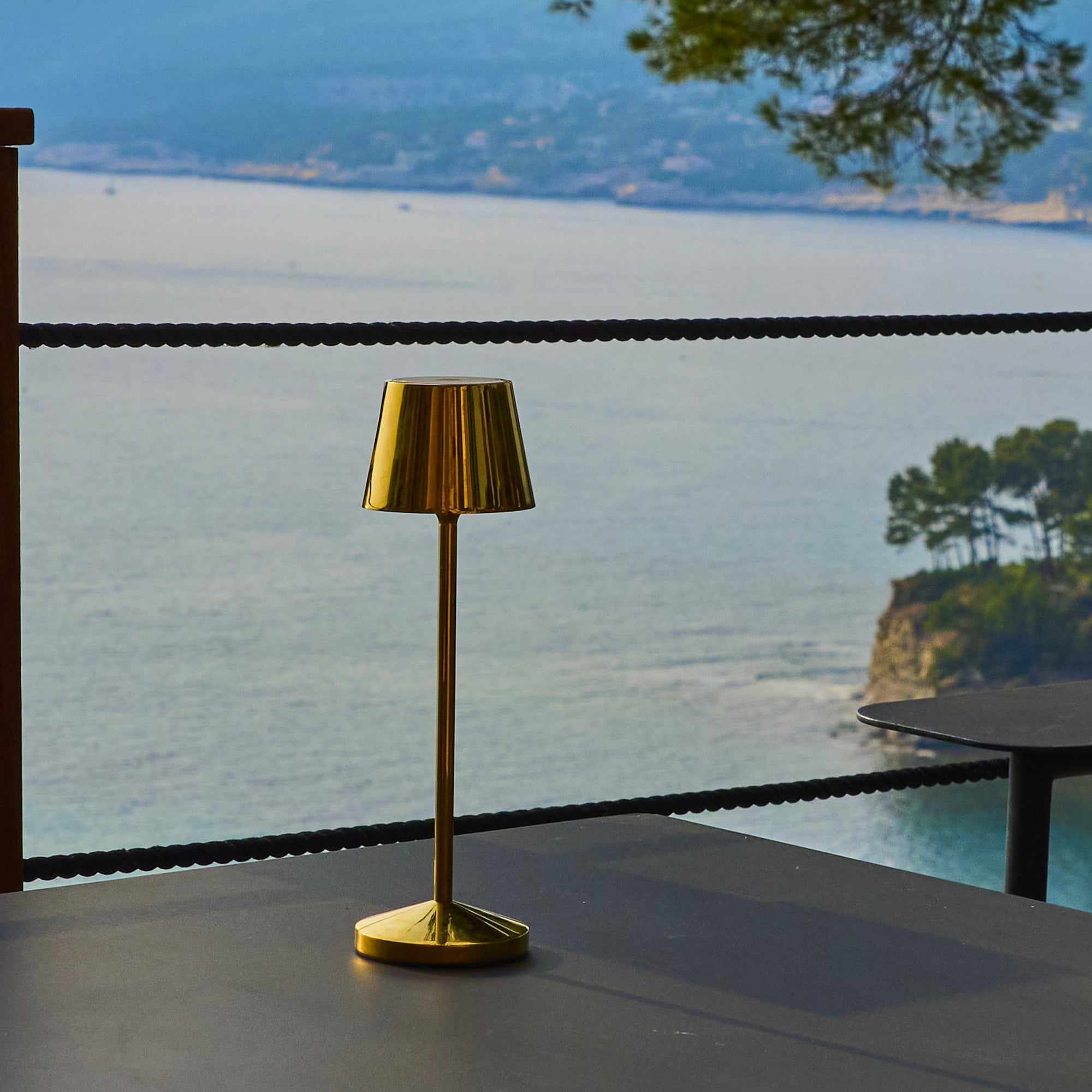 Eine goldene Lampe auf einem Tisch mit Meerblick im Hintergrund. Vor dem Meer befindet sich ein schwarzer Seilzaun.
