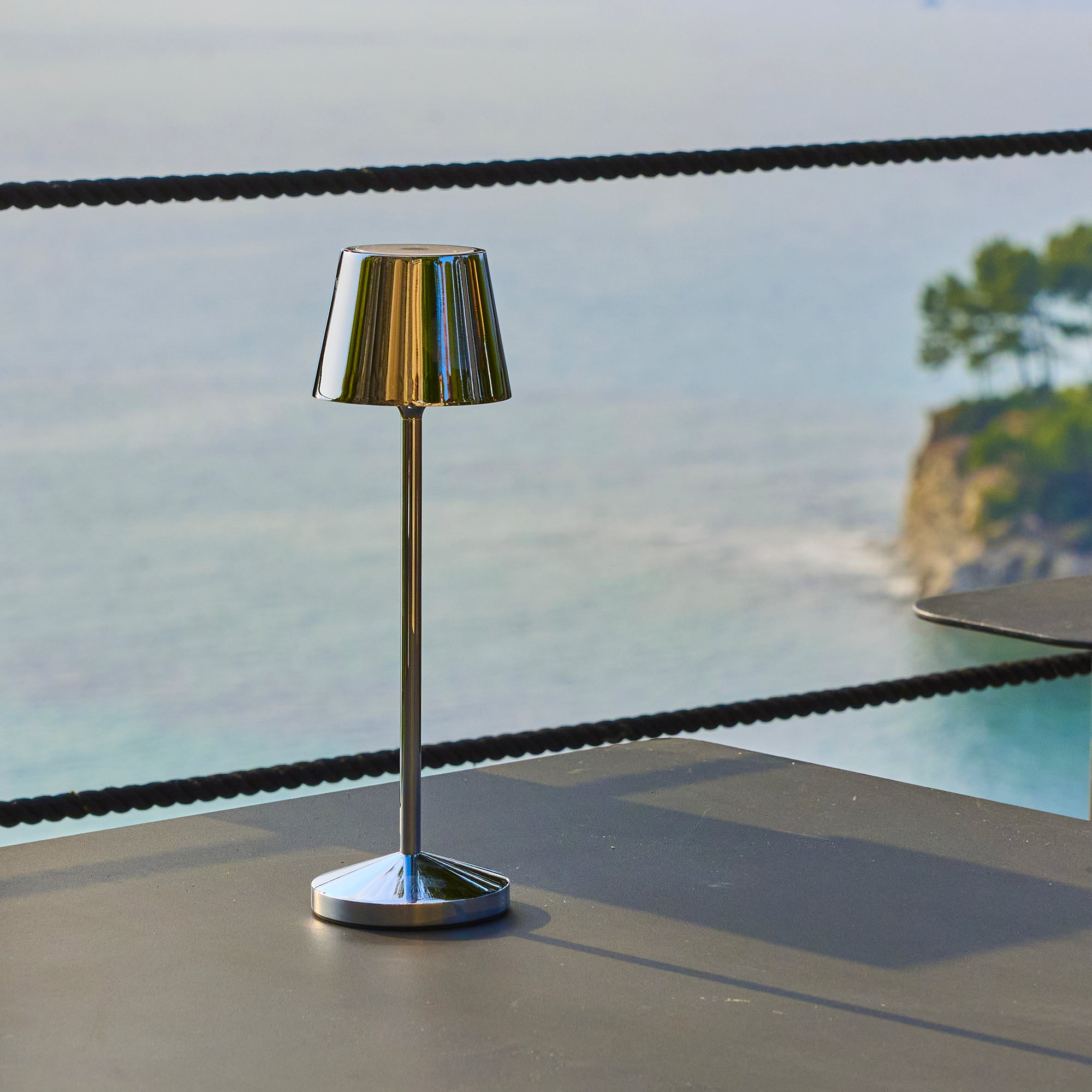 Eine metallische Tischlampe mit reflektierendem Schirm steht auf einem dunklen Tisch mit Blick auf das Meer und eine Insel.